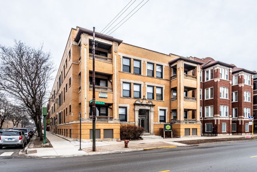 Three-story brick apartment building on a city street. Beige and brown exterior, balconies, overcast sky.