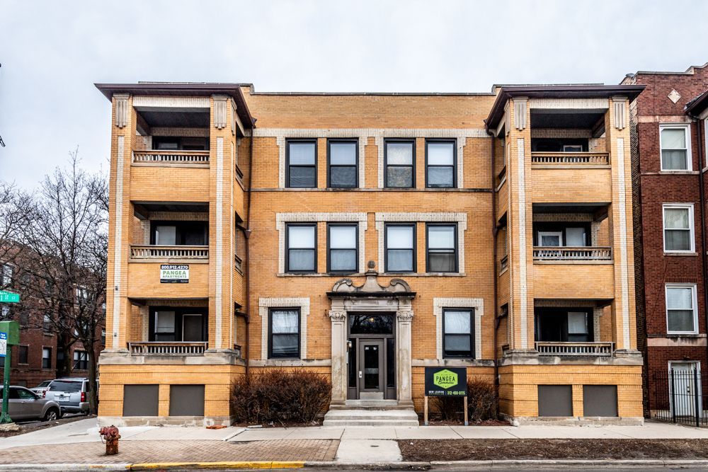 Three-story tan brick apartment building with balconies, dark windows, and a prominent front entrance.