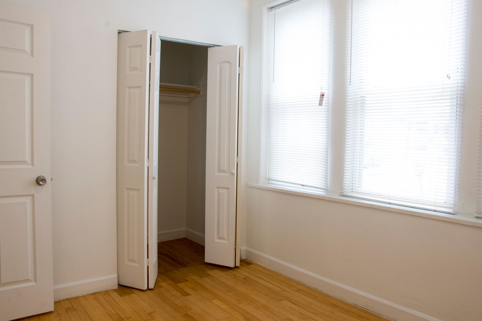 Empty room with a closet and a window with blinds; hardwood floor, white walls and doors.