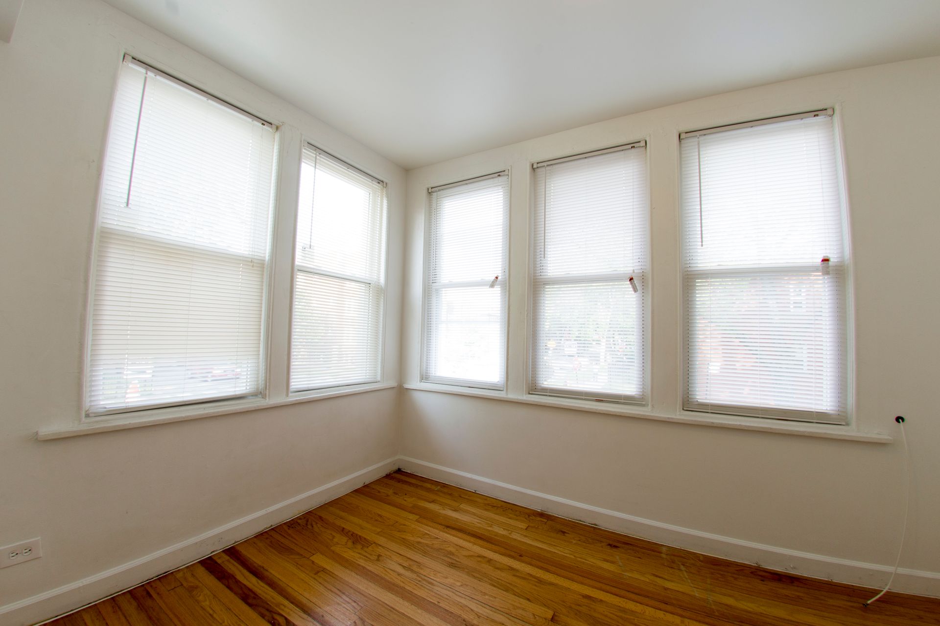 Empty room with wooden floor, four windows with blinds, and white walls.