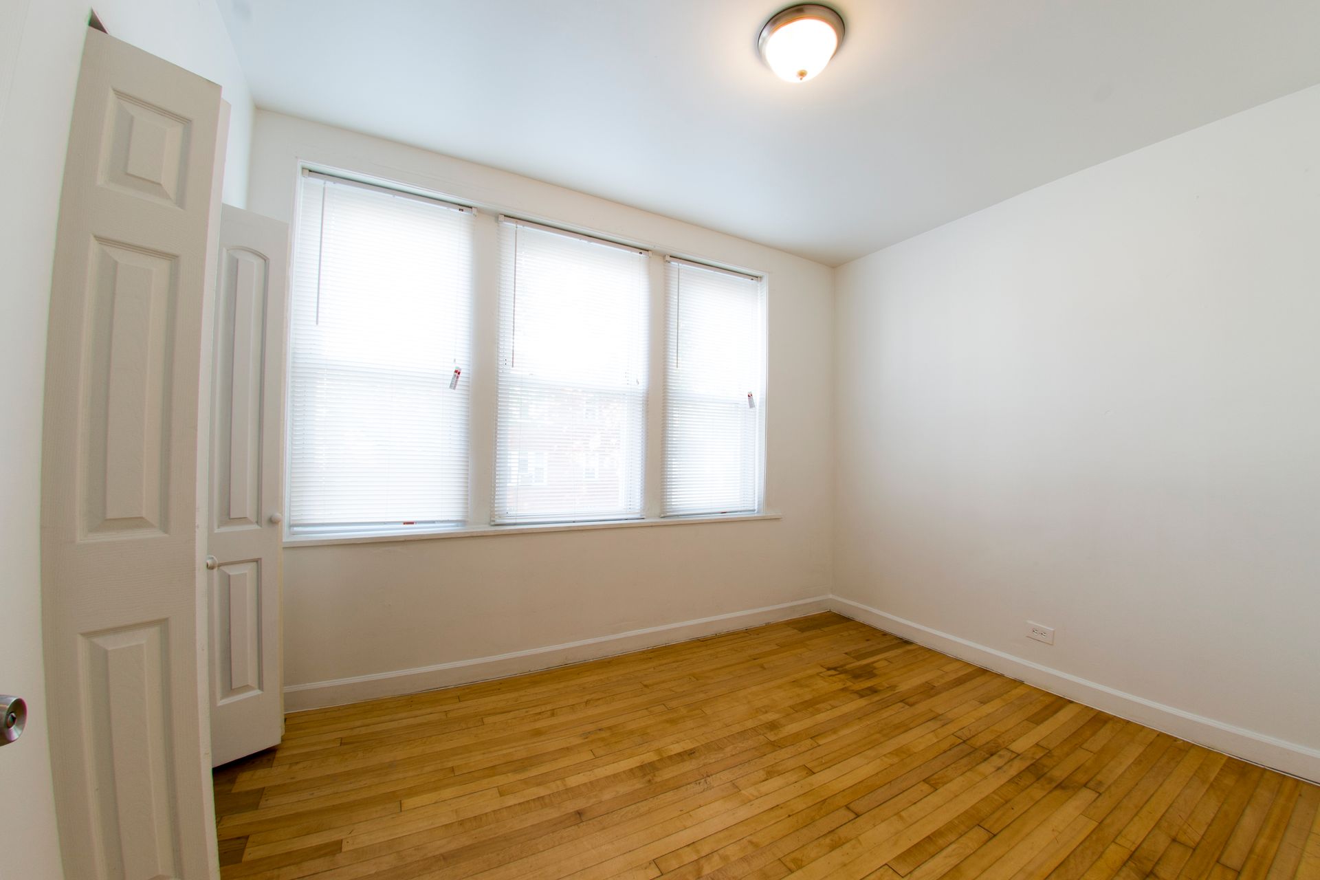 Empty room with hardwood floors, a window with blinds, and a closed door.