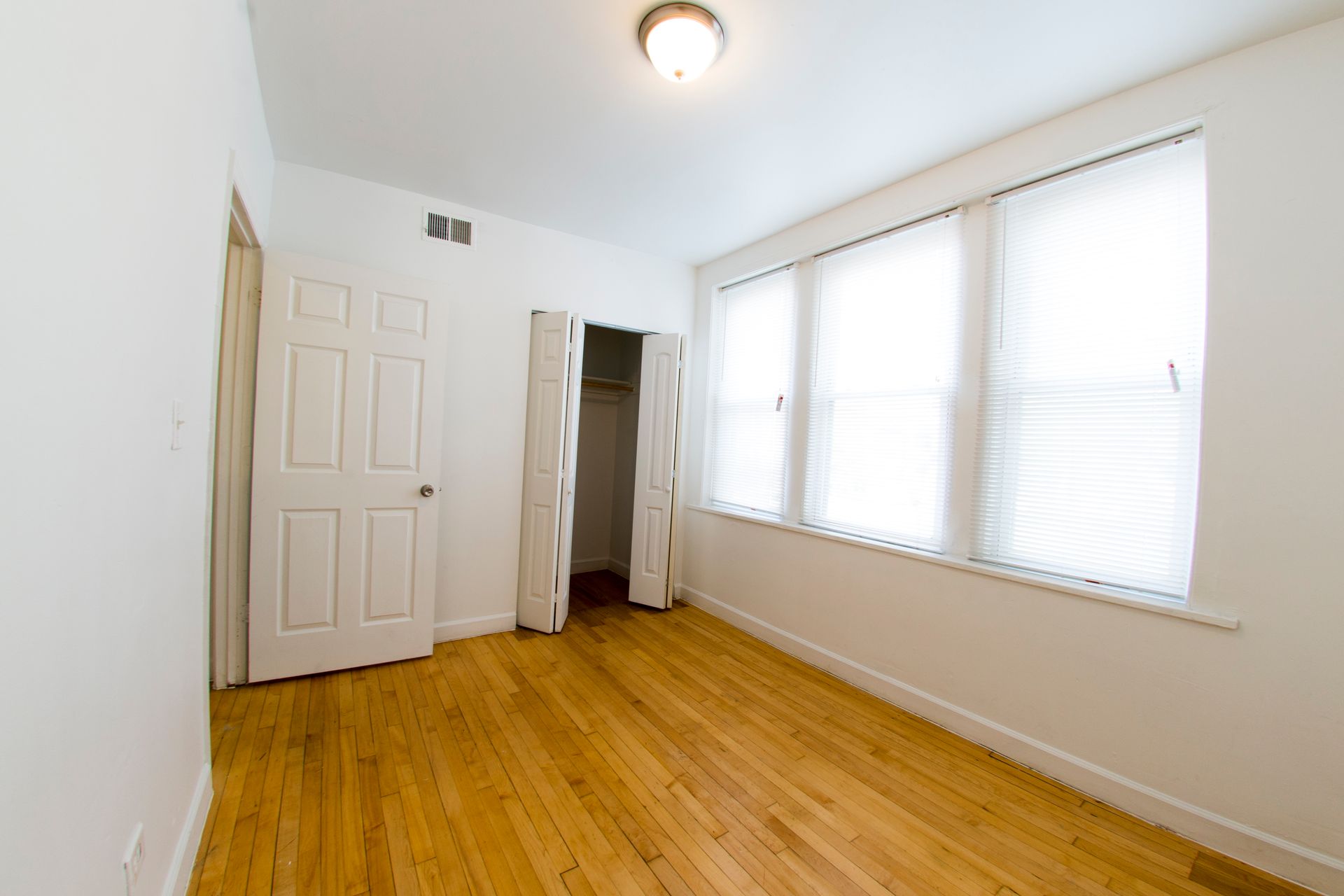 Empty bedroom with wooden floor, white walls, closet, and large window with blinds.