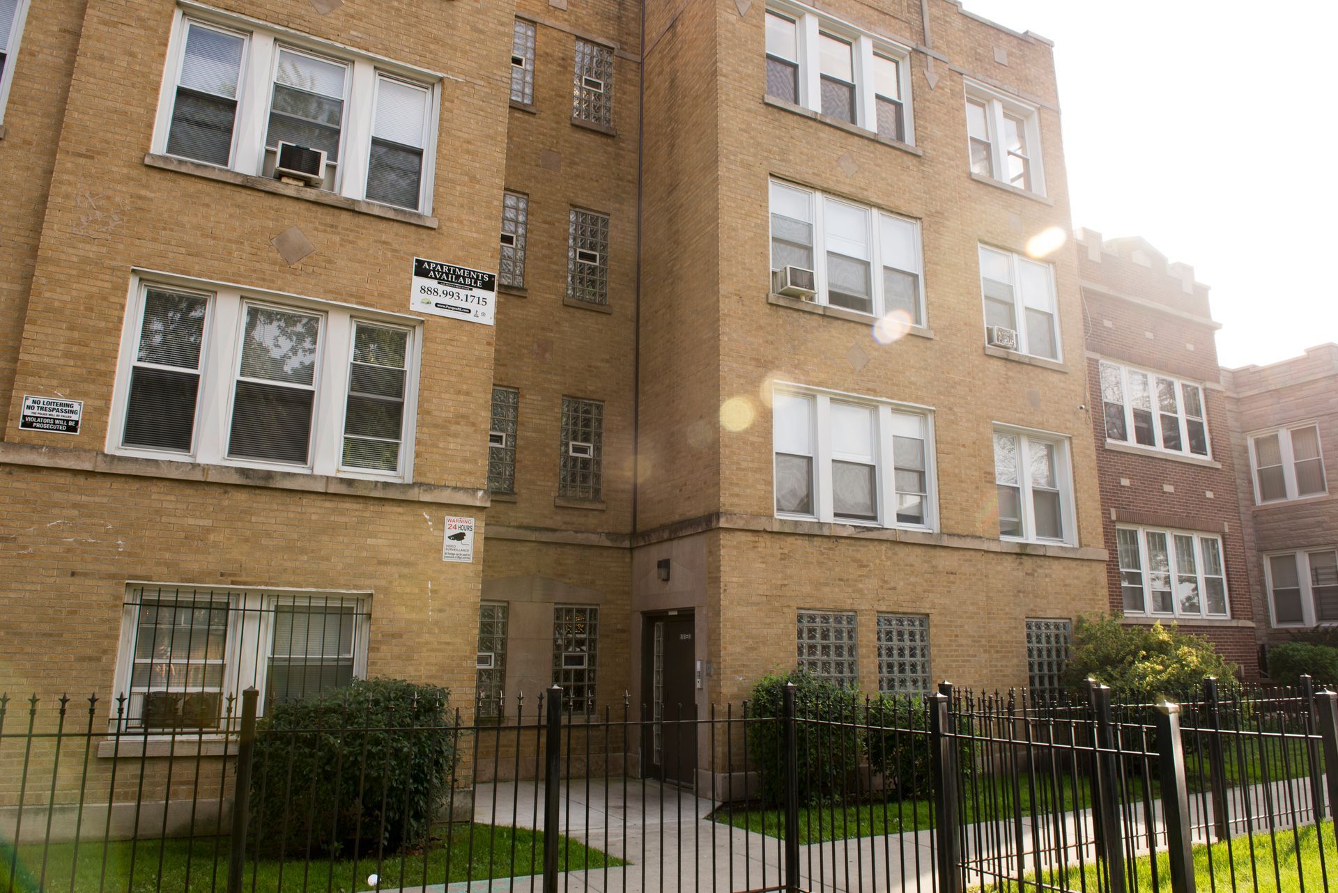 Multi-story brick apartment building behind a wrought iron fence, sunny day.