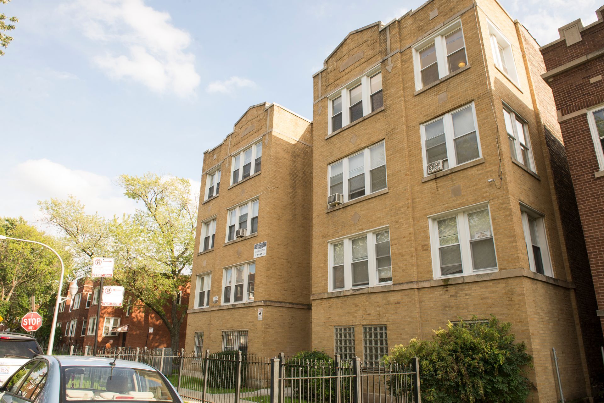 Brick apartment buildings with multiple windows against a cloudy sky. A car and bushes are in the foreground.