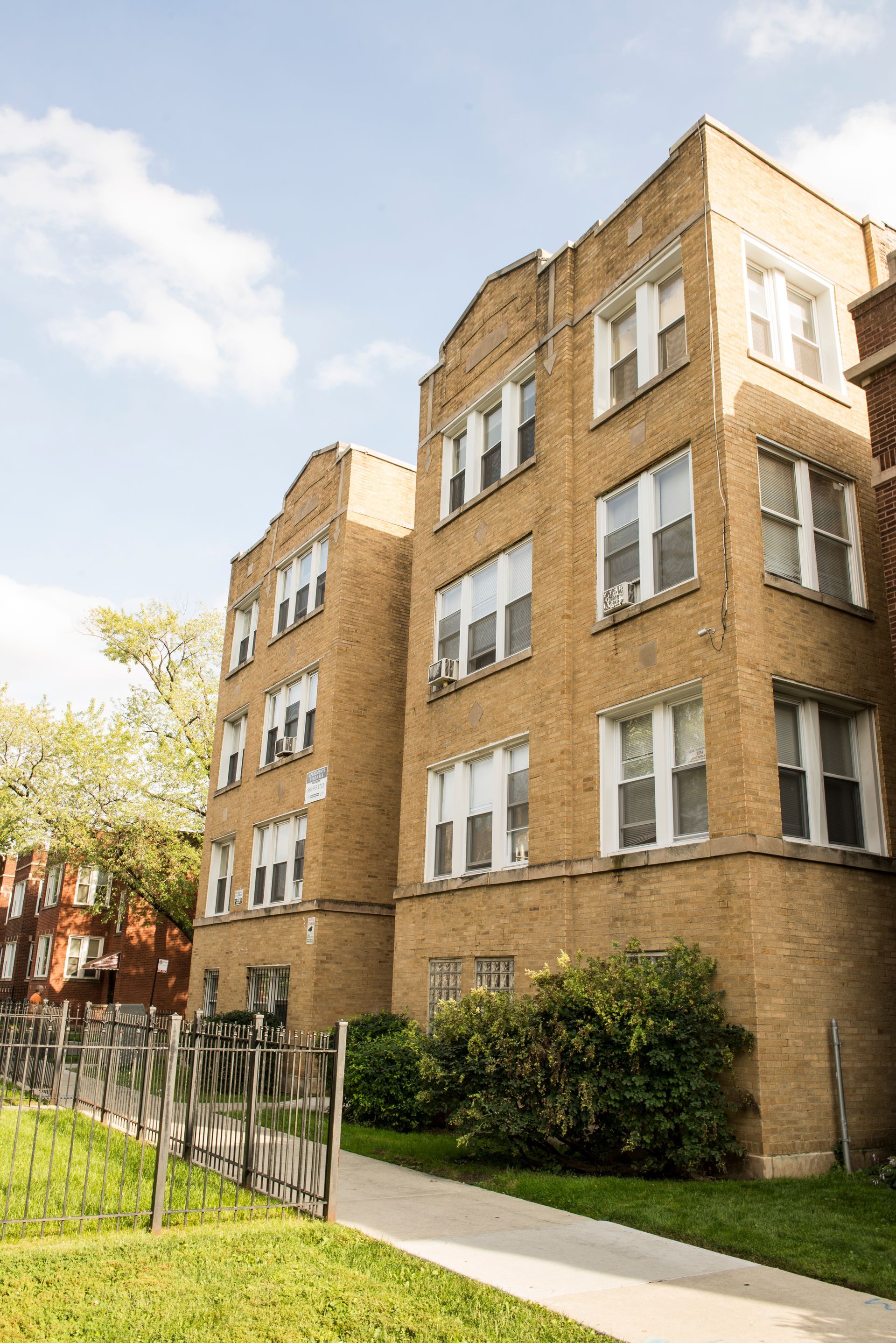 Multi-story brick apartment building on a sunny day with a walkway and grassy area.