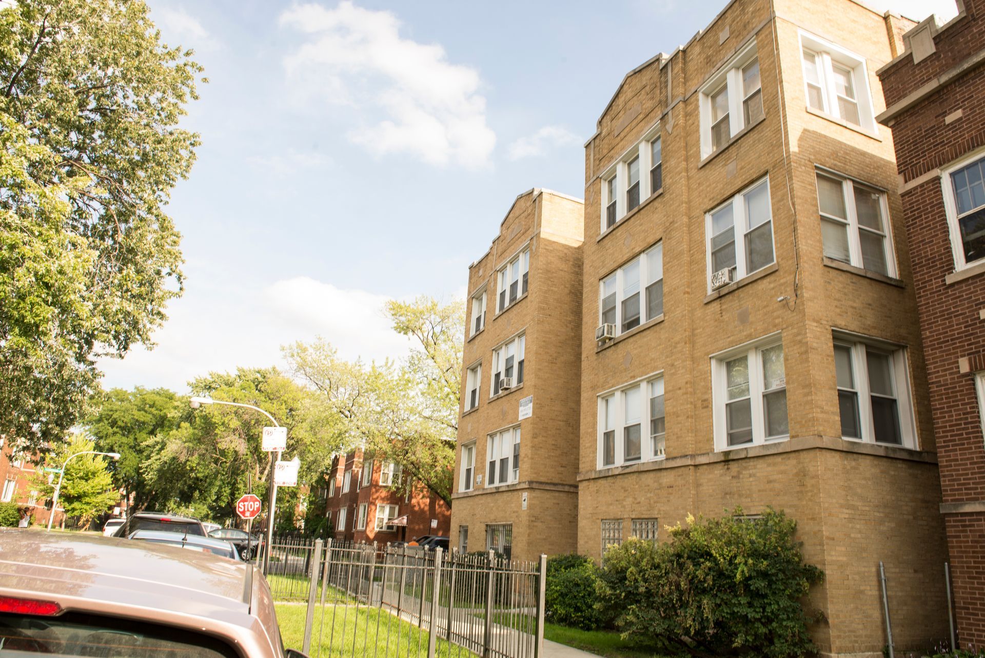 Tan brick apartment building on a sunny street, partial view of a car and trees.
