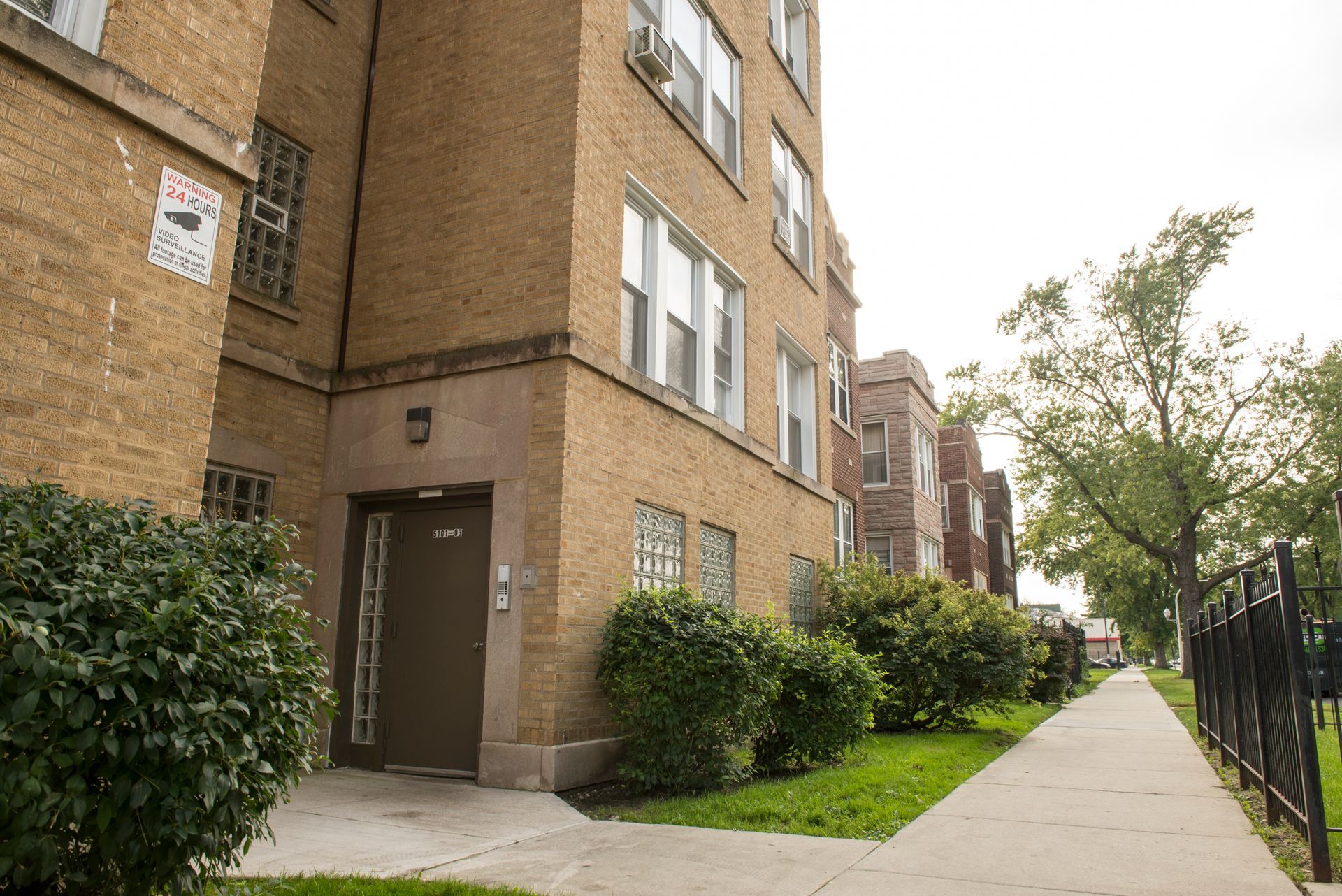 Brown brick apartment building with a sidewalk and green shrubbery.