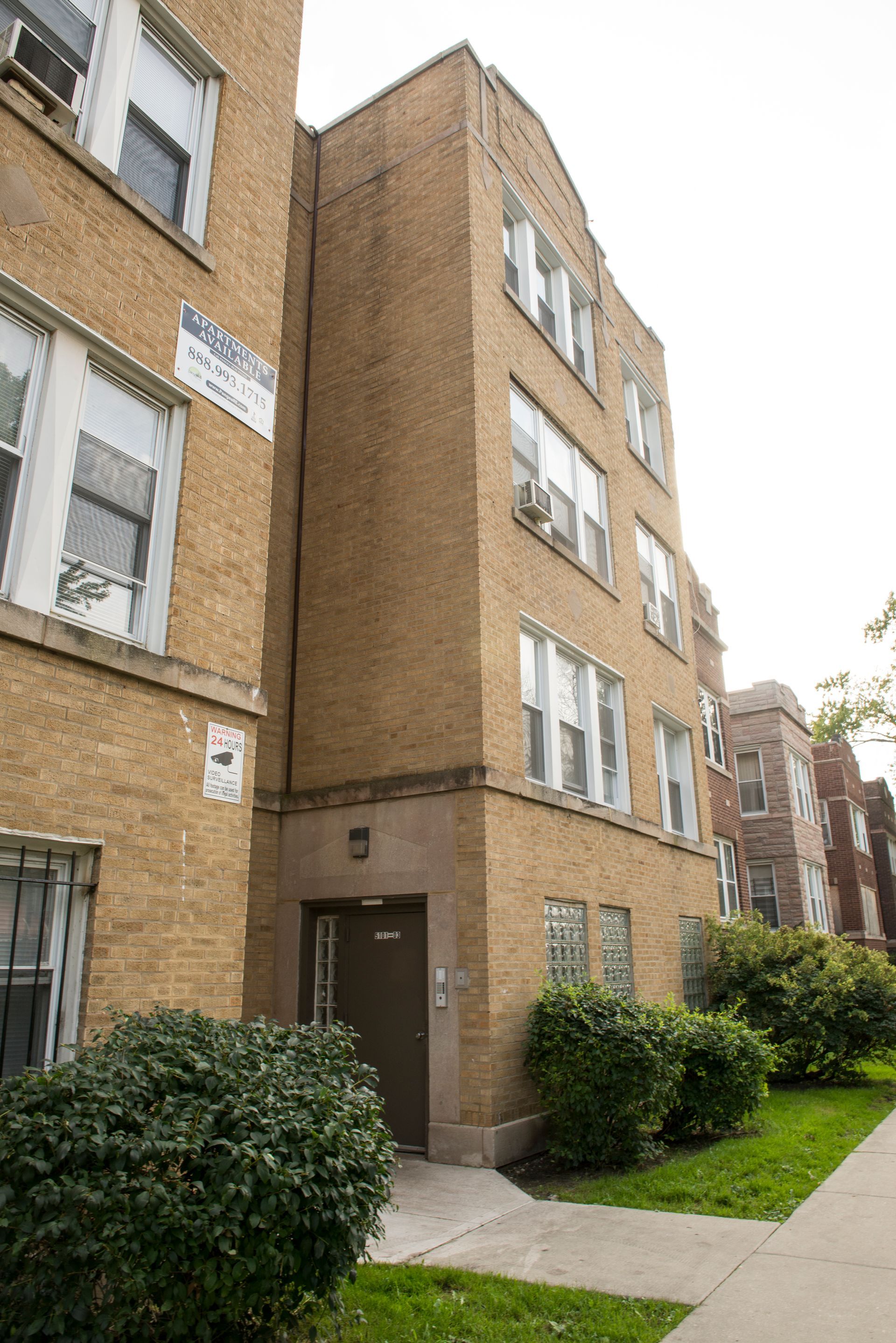 Multi-story brick apartment building with entrance and windows, on a green lawn.