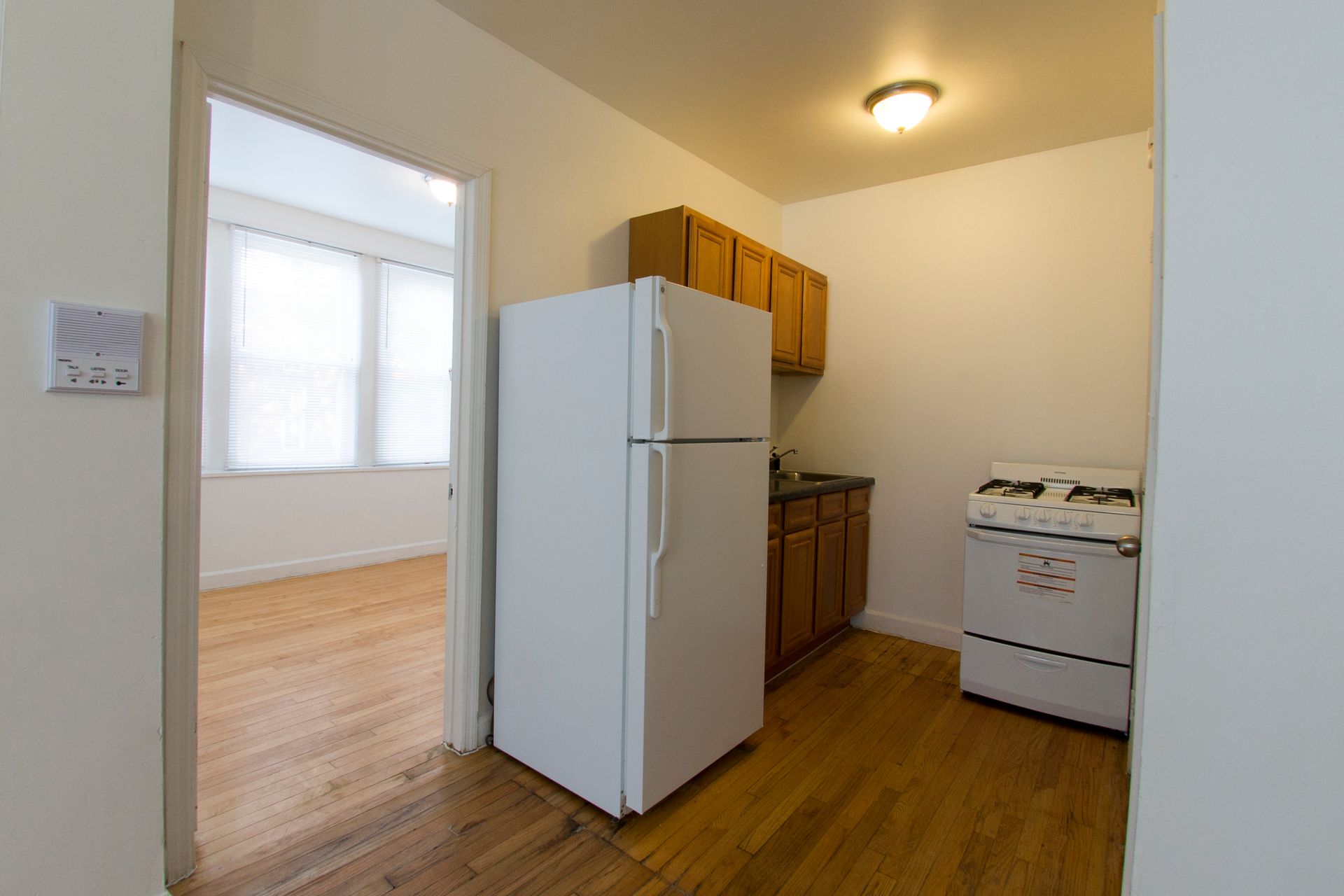 Small kitchen with a refrigerator, stove, and cabinets; doorway to a room with windows and wood flooring.