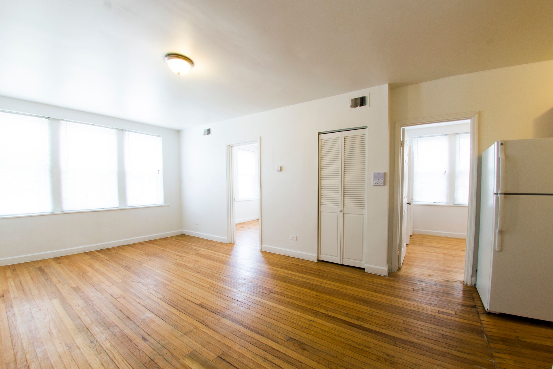 Empty apartment interior with wood floors, large windows, white walls, and a refrigerator.