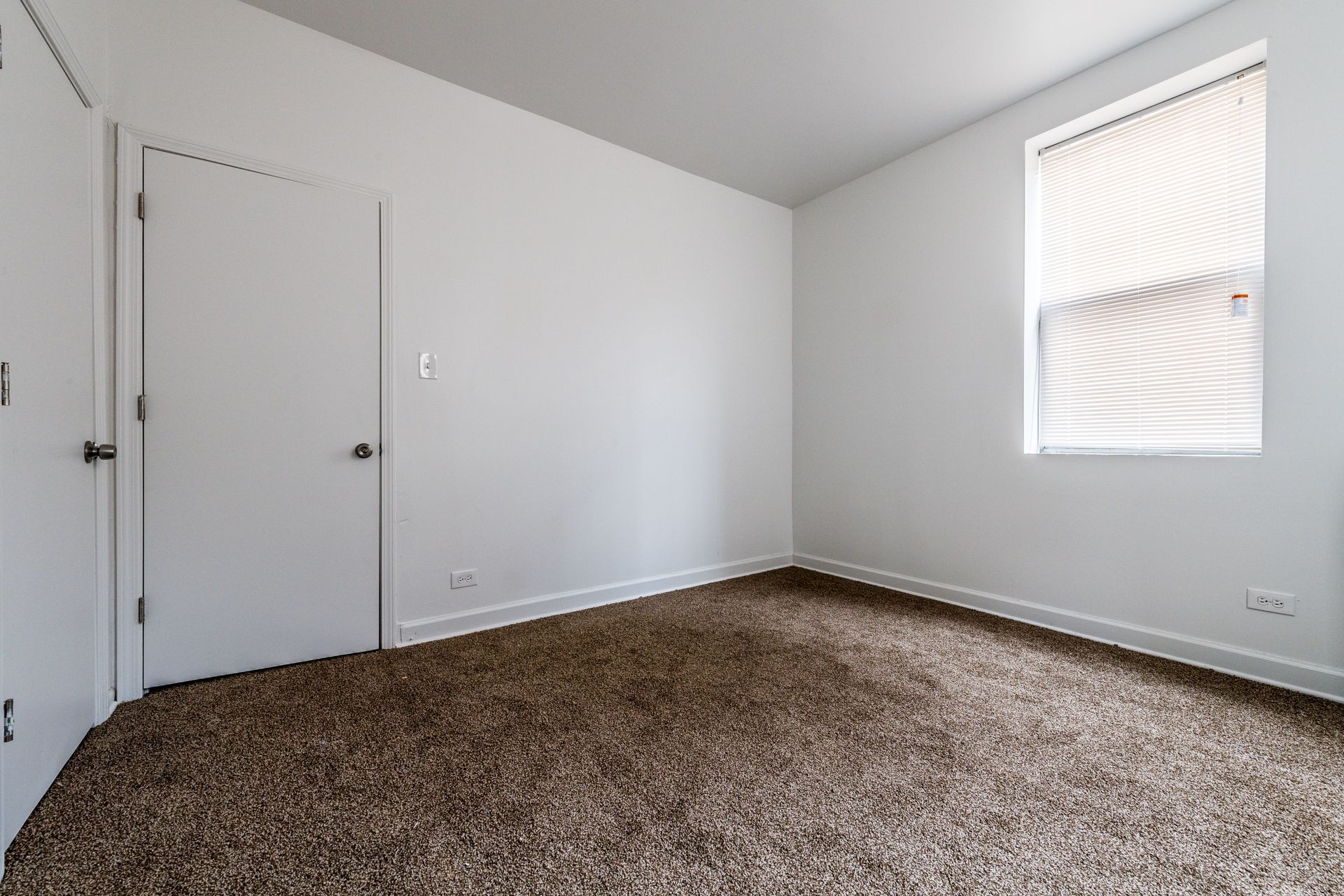 Empty room with brown carpet, white walls, closed door, and window with blinds.