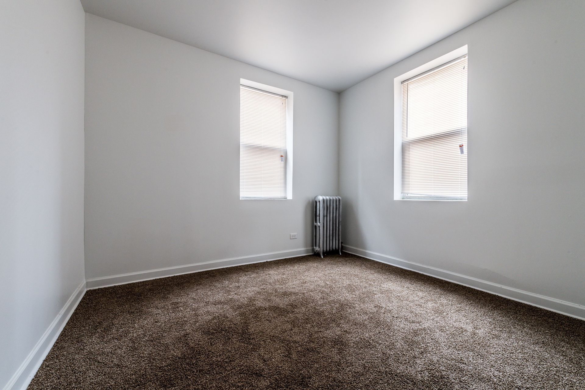 Empty room with beige carpet, white walls, two windows with blinds, and a radiator.