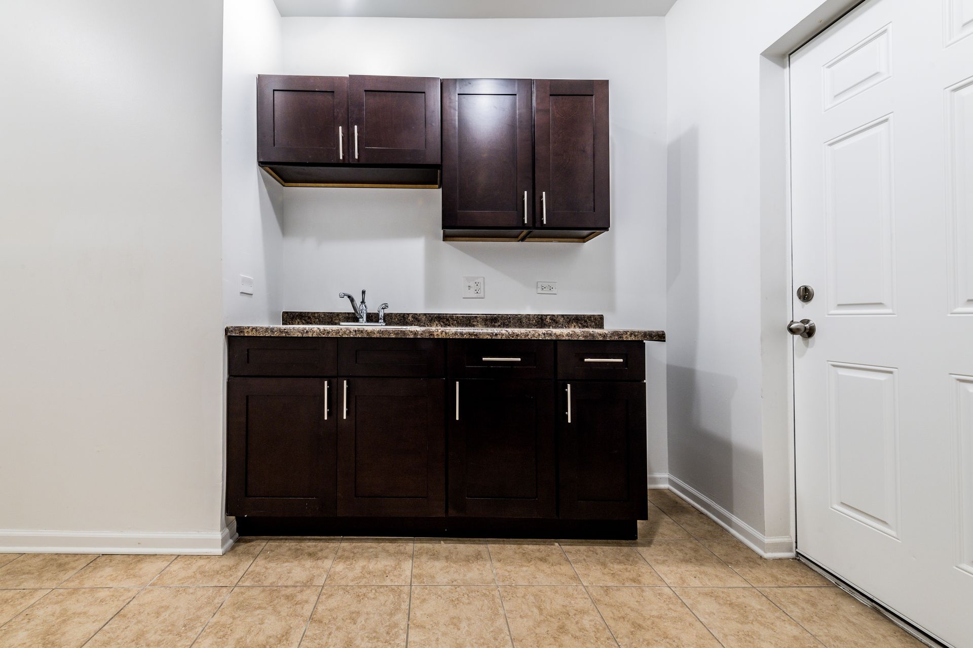 Dark brown cabinets with sink and white door in room with tiled floor.