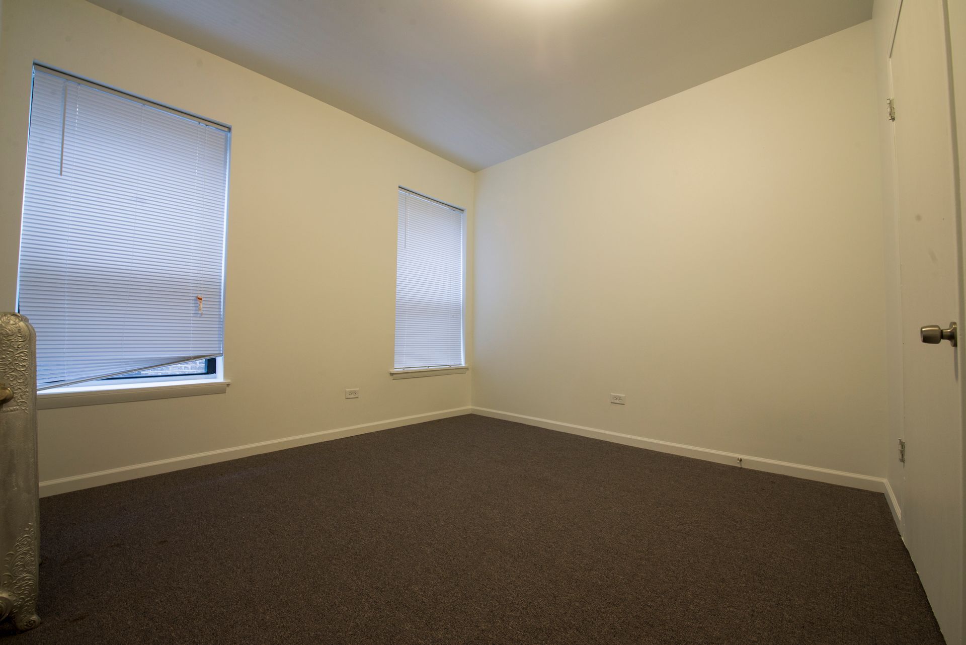 Empty room with brown carpet, two windows with blinds, and a white door.