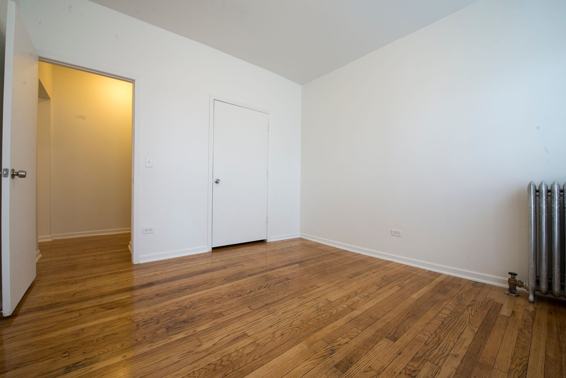 Empty room with hardwood floors, white walls, two white doors, and a radiator.