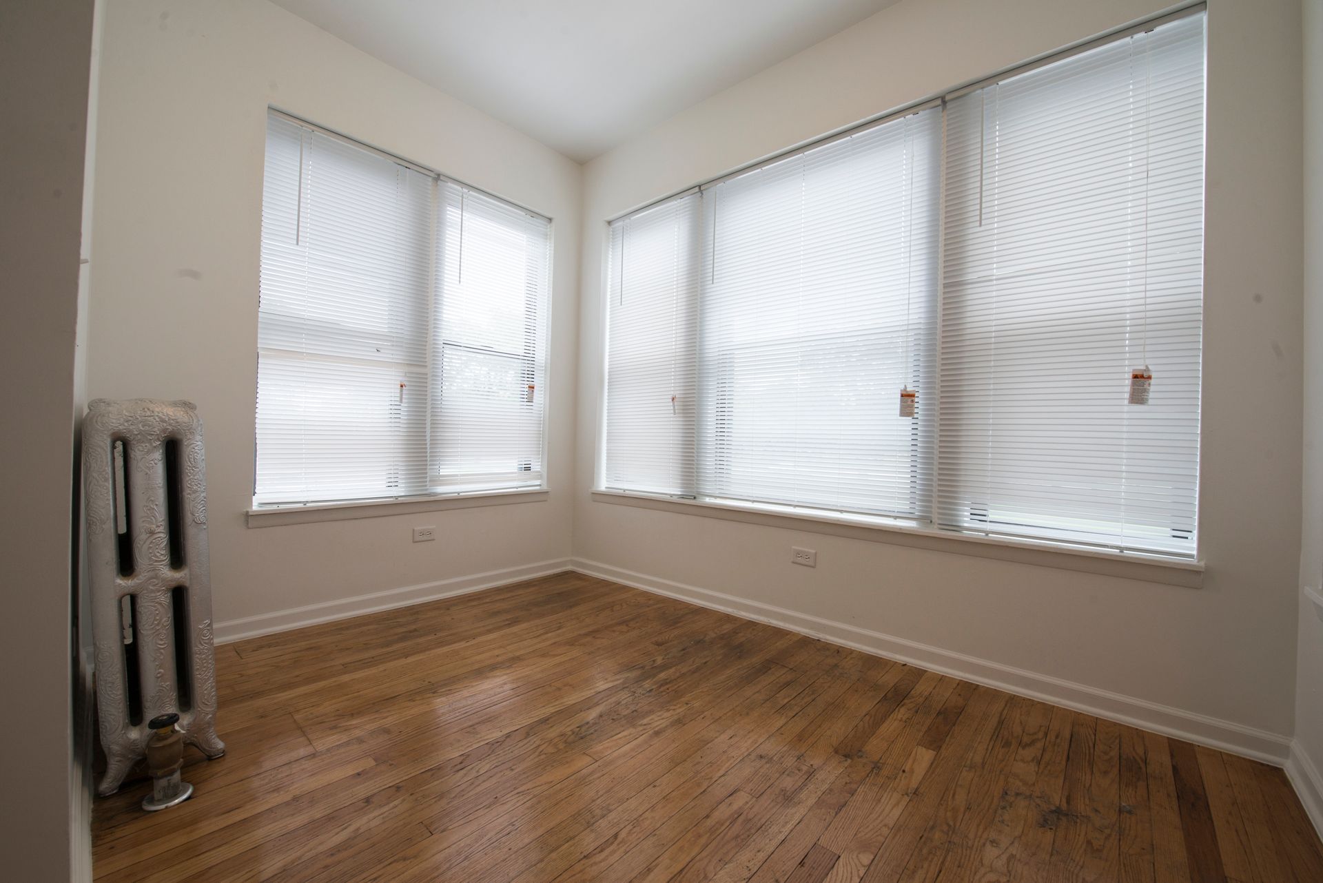 Empty room with hardwood floors, three large windows with blinds, and a radiator.
