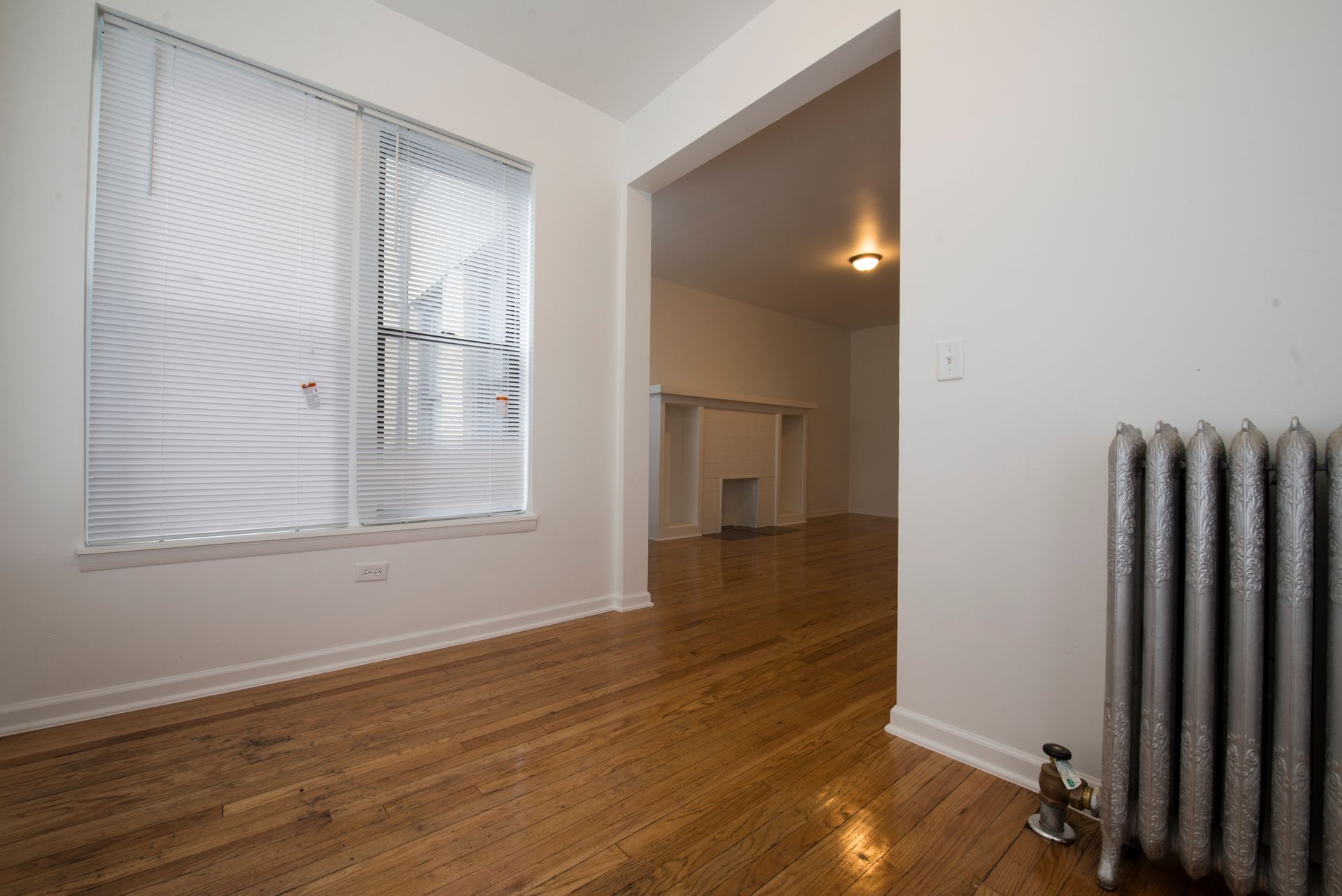 Empty room with hardwood floors, a radiator, and a view into another room with a fireplace.
