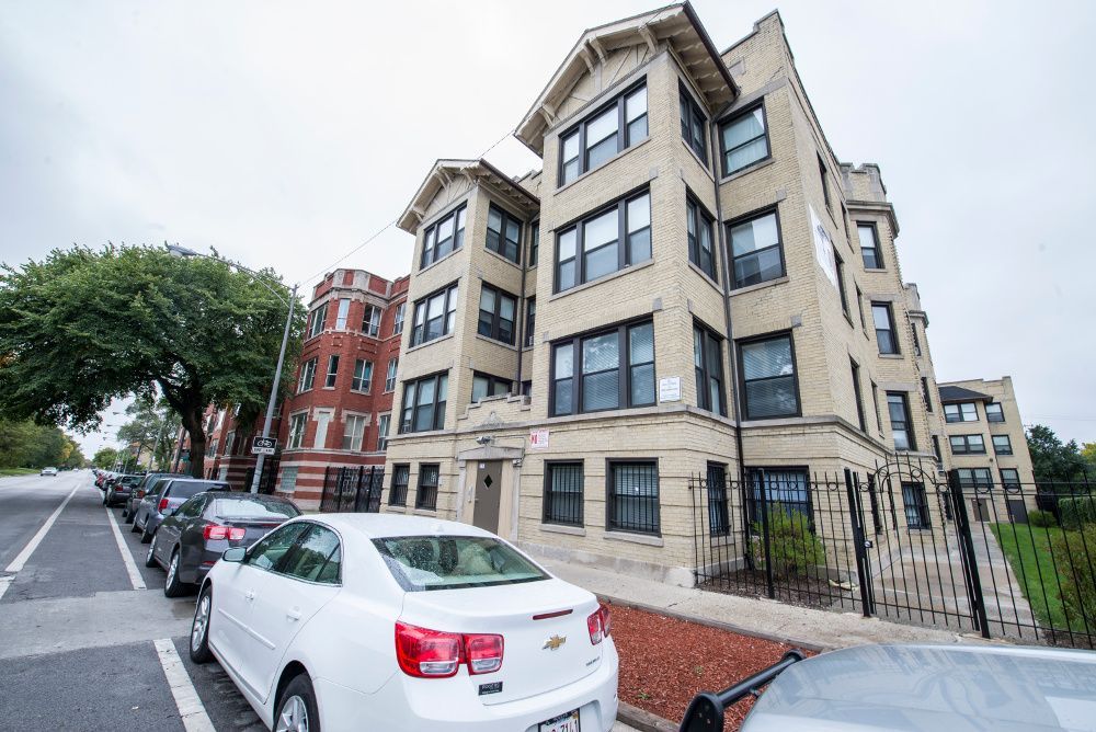 Multi-story brick apartment building on a city street, cars parked in front, overcast day.