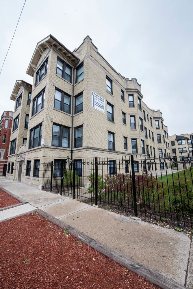 Multi-story brick apartment building behind a black metal fence and sidewalk on a cloudy day.
