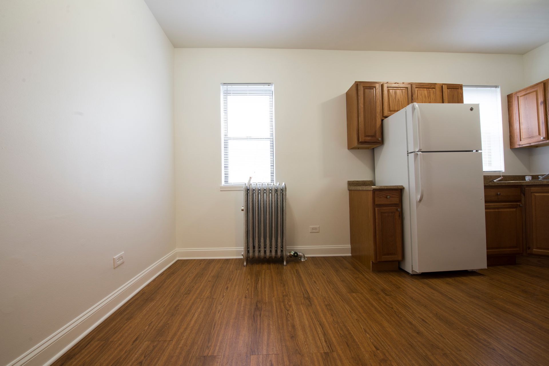Empty kitchen with wooden cabinets, refrigerator, and radiator against a white wall and wood flooring.