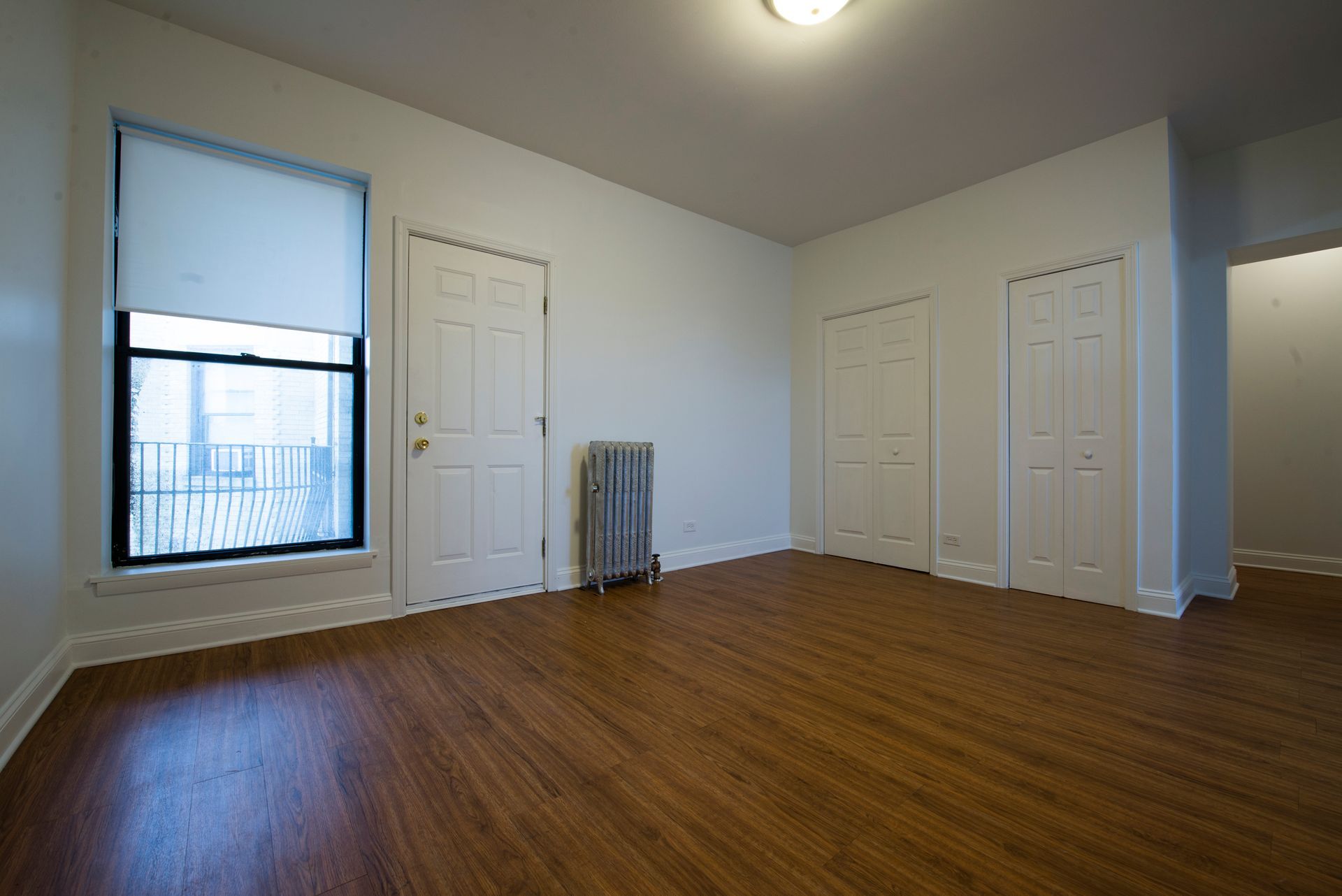 Empty room with hardwood floors, a window with a shade, and white doors.