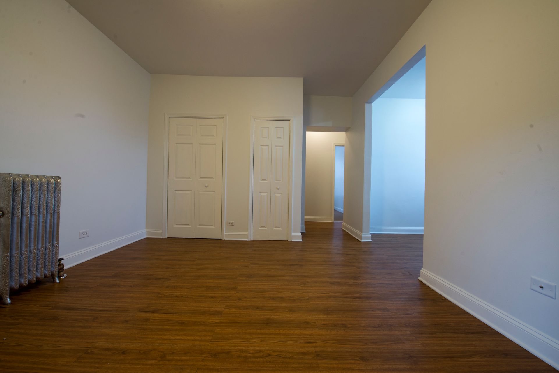 Empty room with hardwood floors, white walls, two closets, and a hallway.