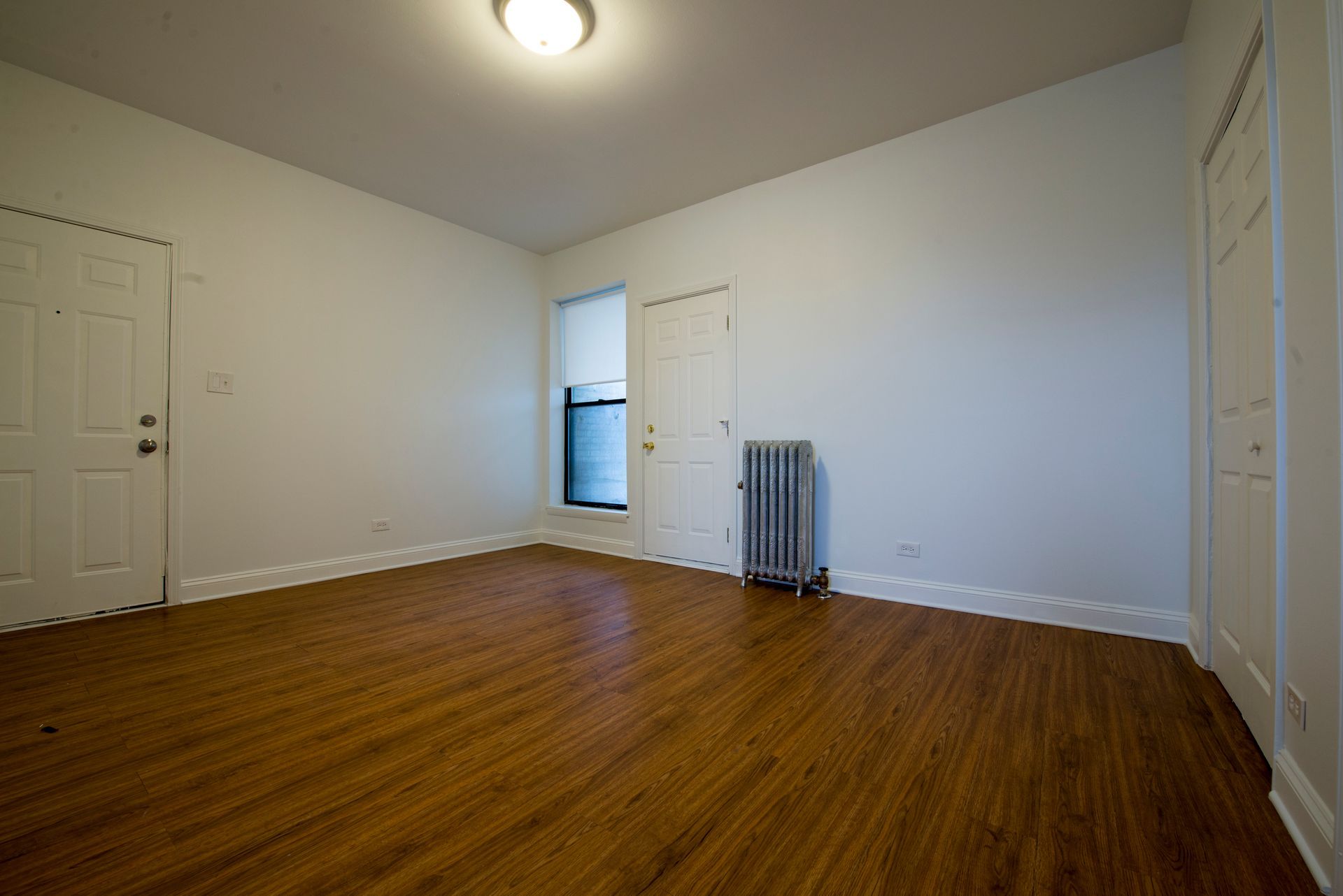 Empty room with hardwood floors, white walls, and a radiator. Includes a door, window, and overhead light.