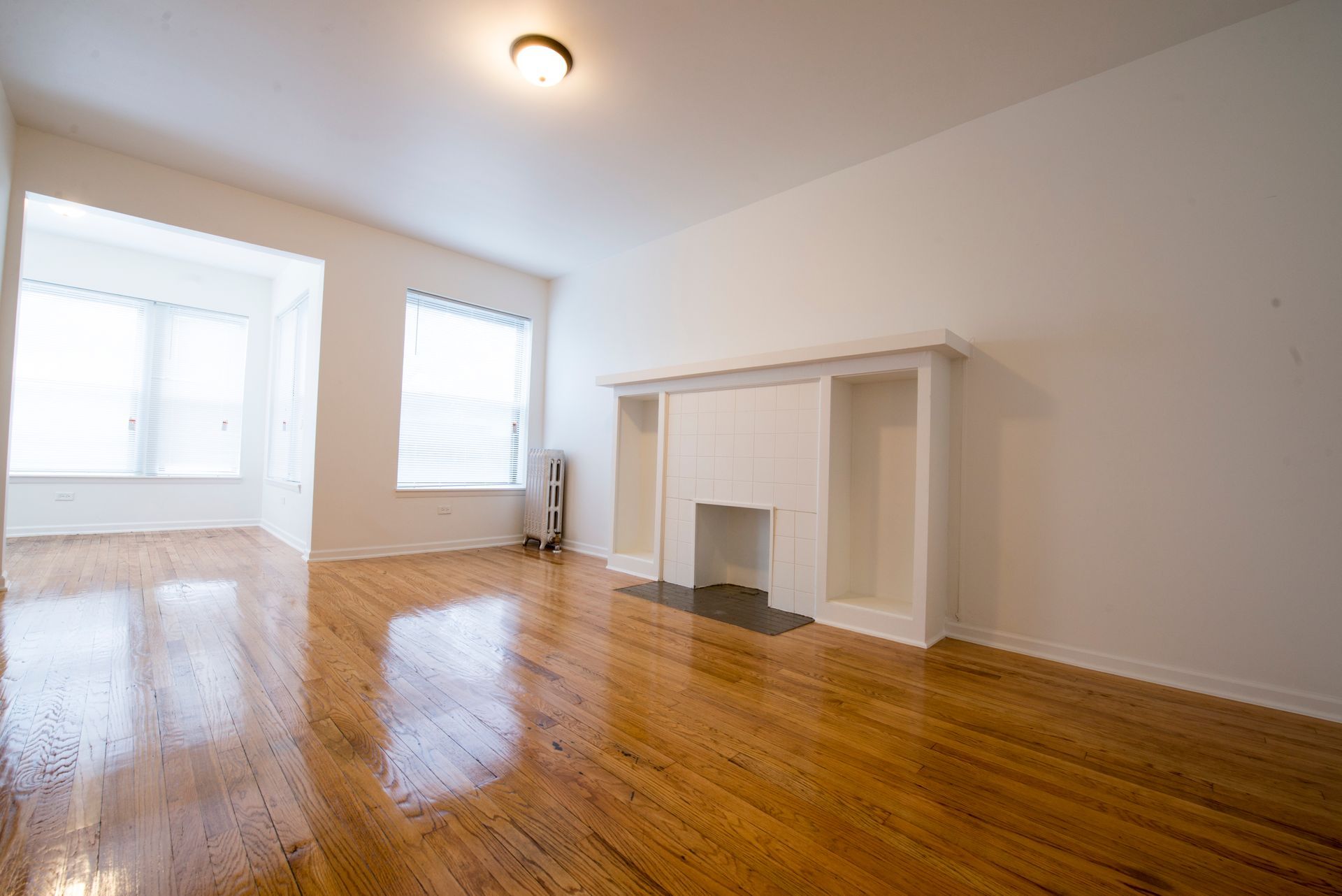 Empty room with hardwood floors, white walls, and fireplace. Sunlight streams through windows.