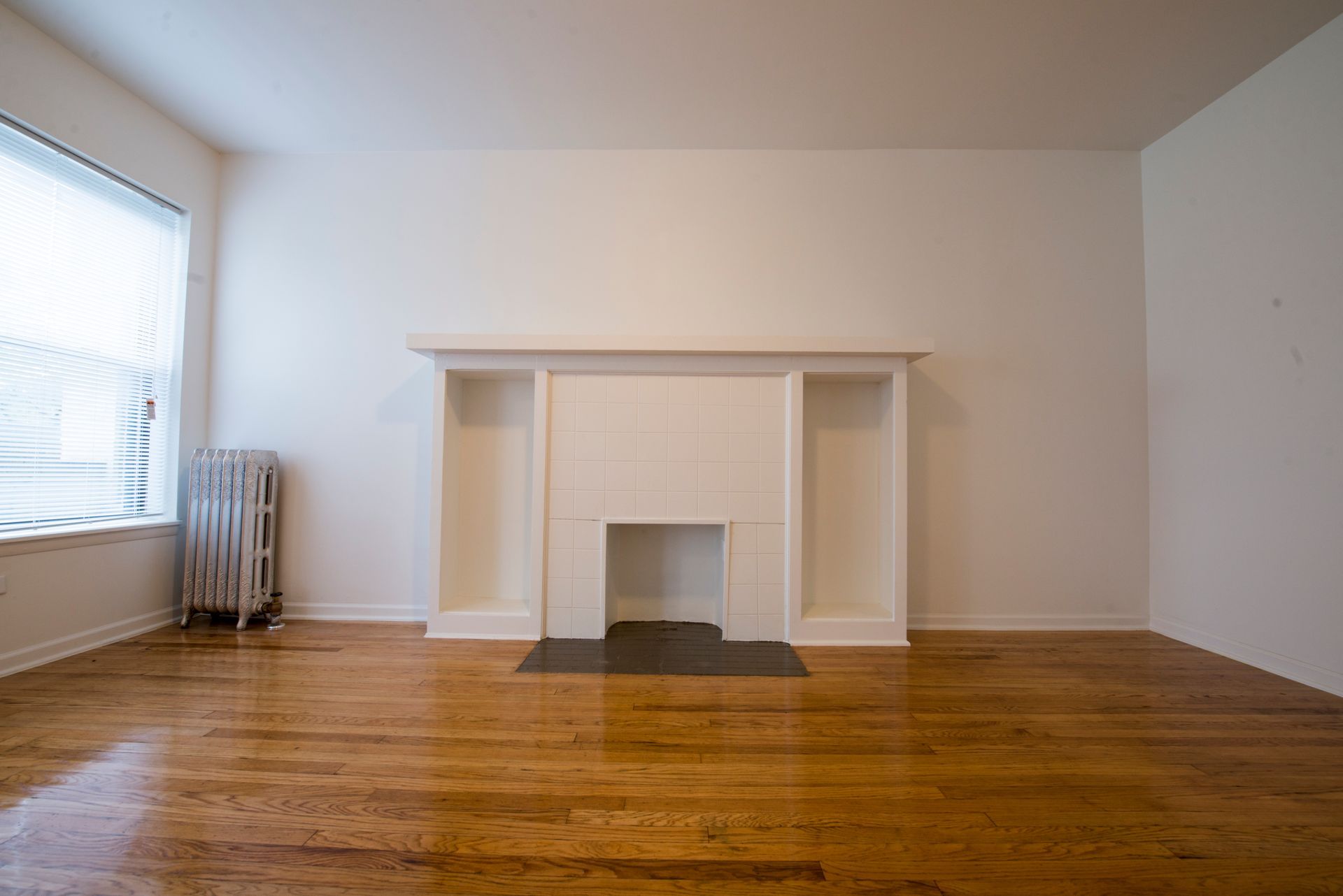 Empty room with a white fireplace and built-in shelving, hardwood floor, radiator, and window with blinds.
