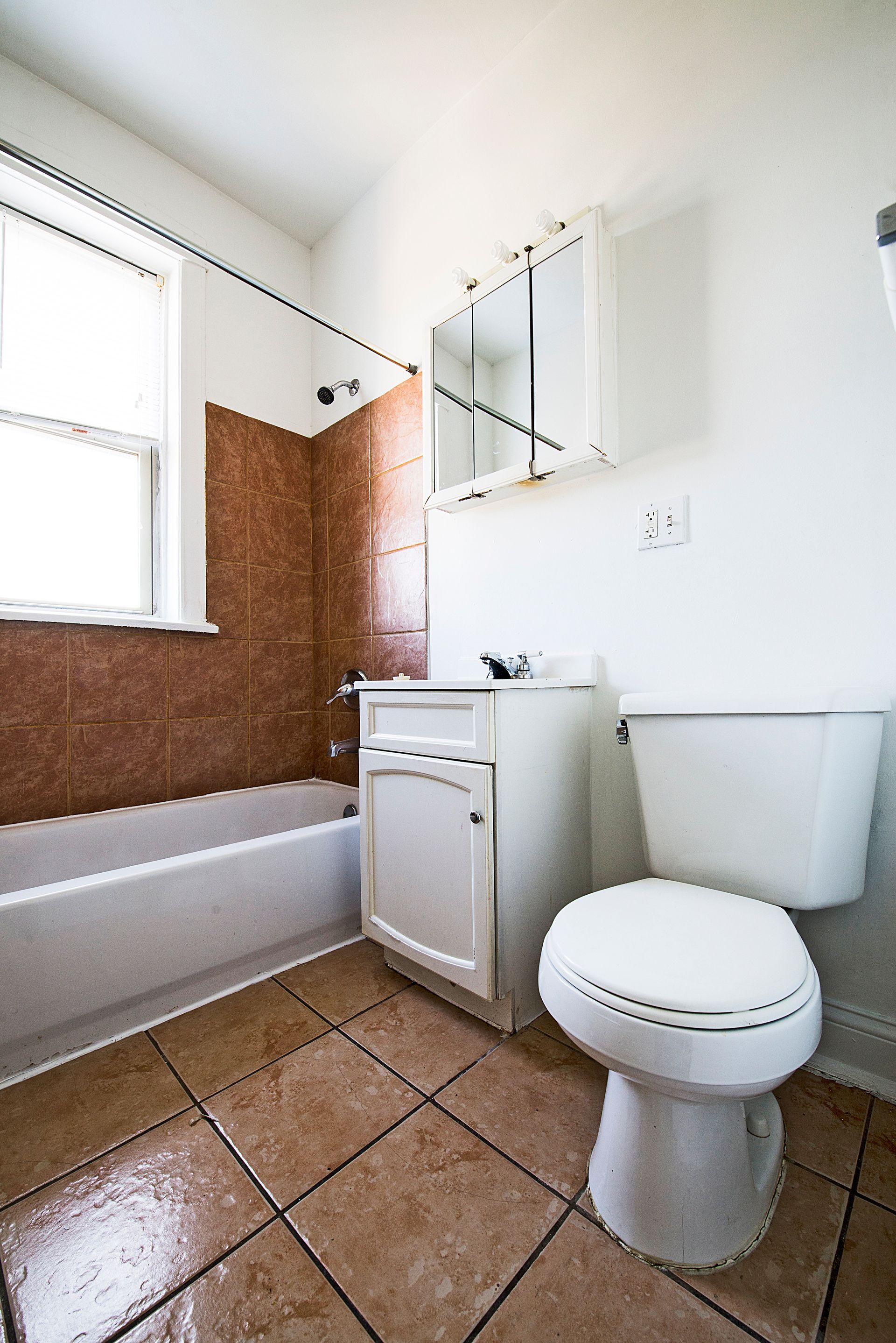 Bathroom with white fixtures, brown tiled floor, white vanity, and tub with brown tiled wall.