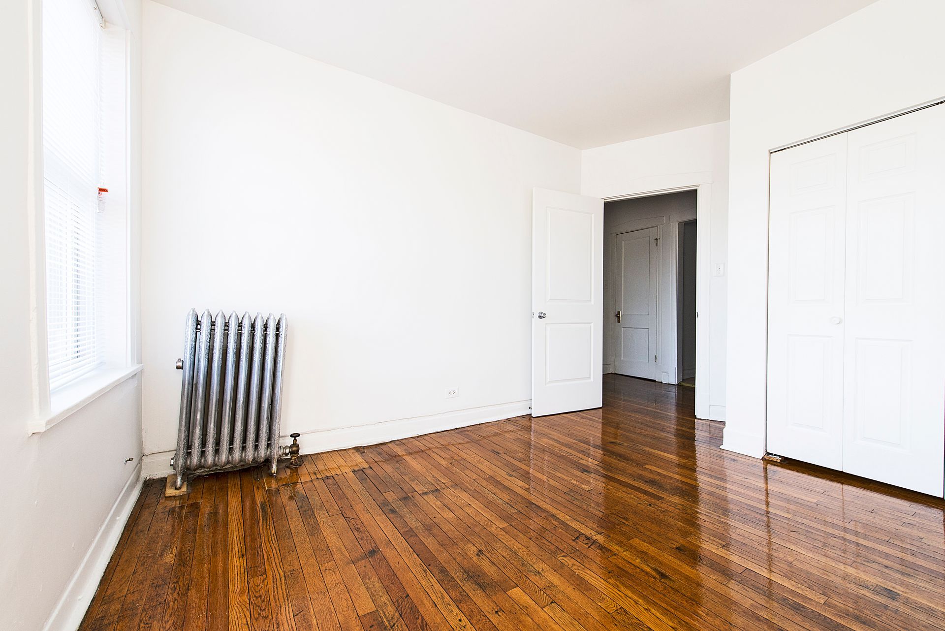 Empty room with hardwood floors, radiator, and white walls. Doorway leads to a hallway.