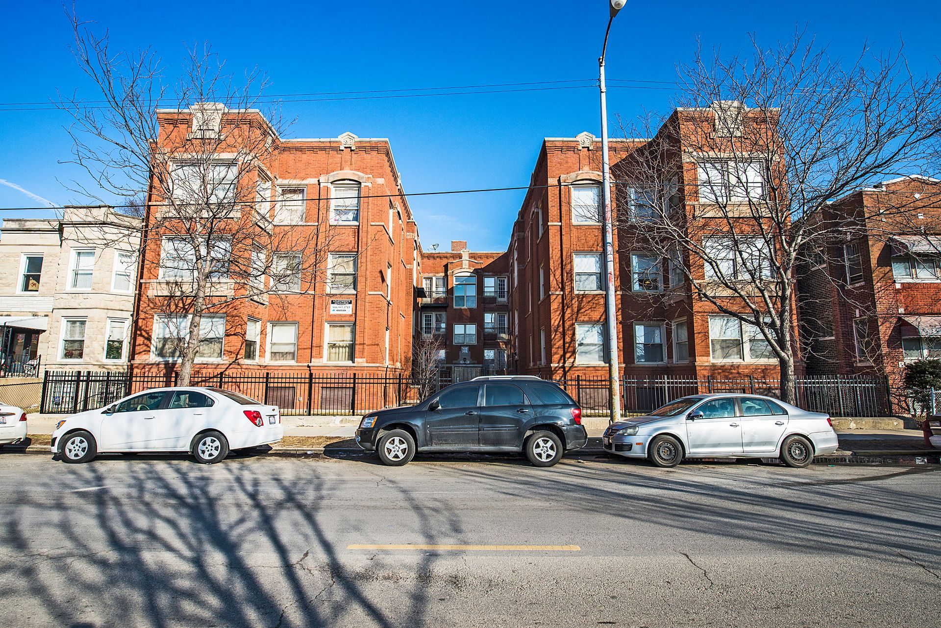 Three-story brick apartment buildings on a city street with parked cars. Clear blue sky overhead.