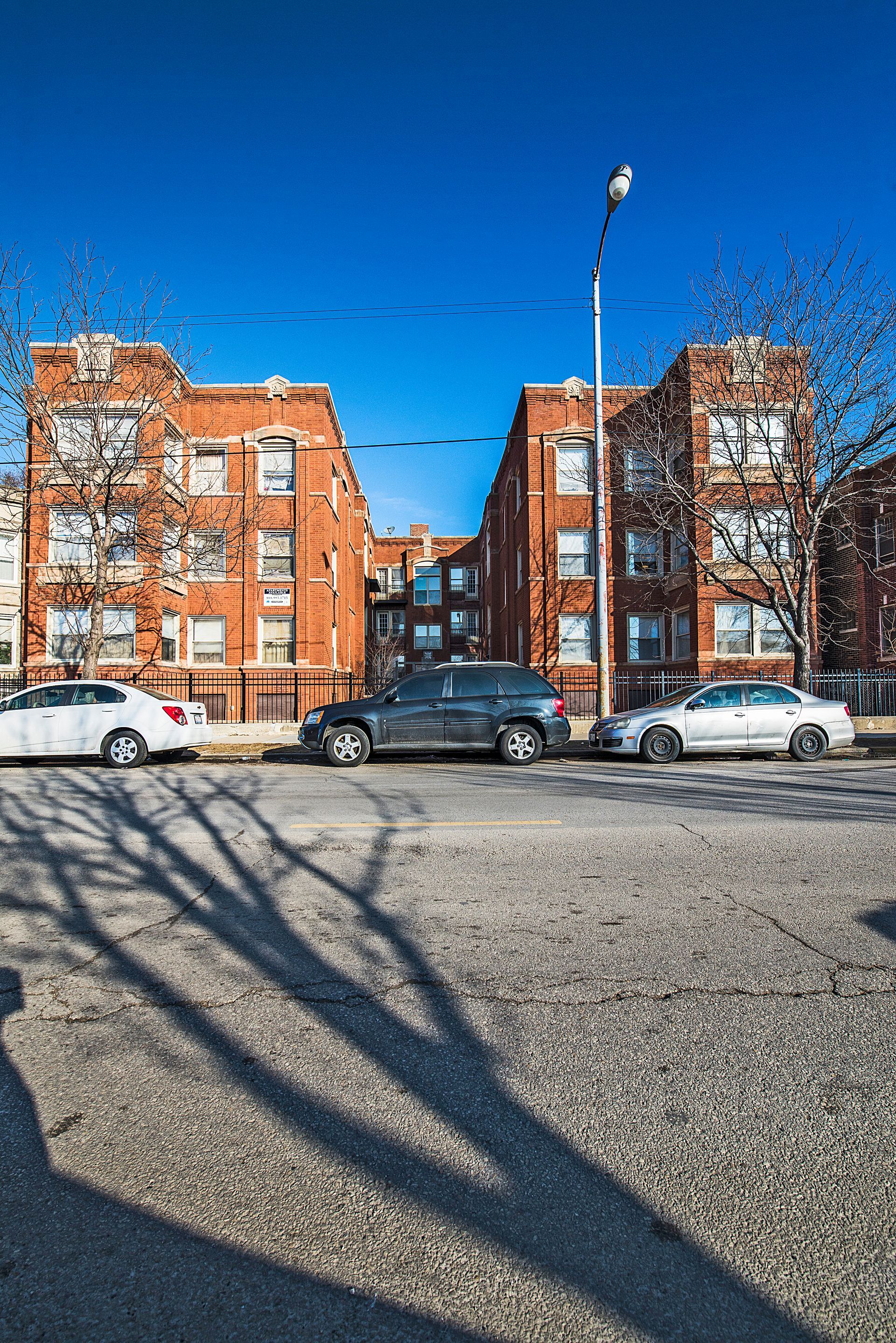 Brick apartment buildings with parked cars on a sunny day. Tree shadow on the gravel ground.