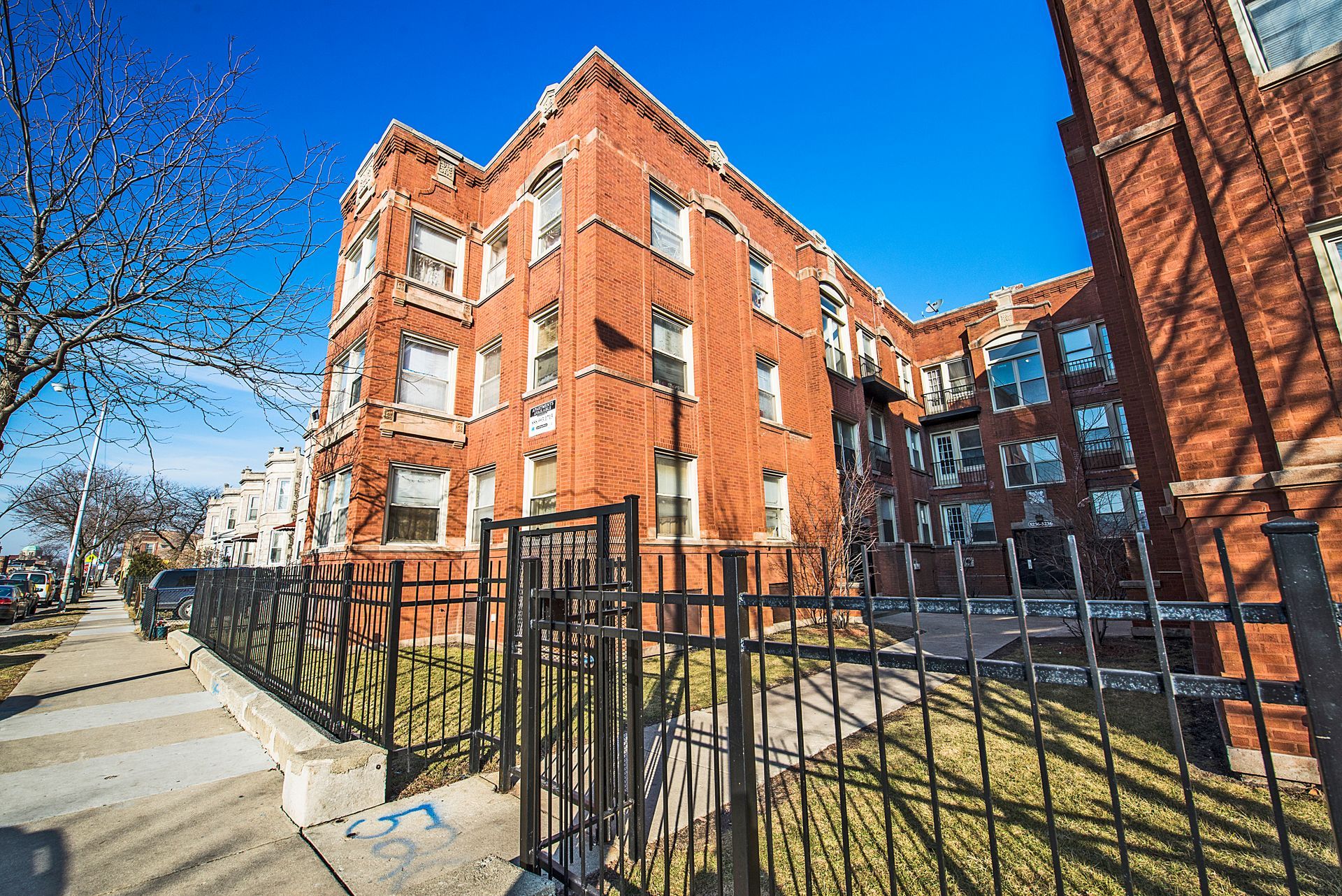 Red brick apartment building behind a black metal fence on a sunny day.