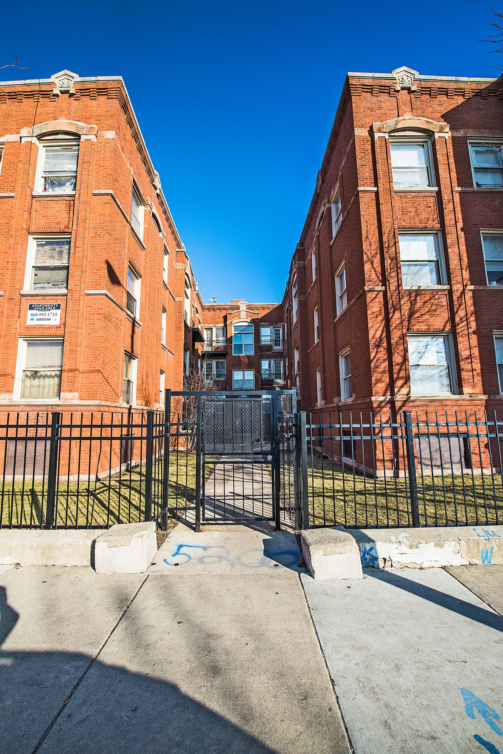 Two brick apartment buildings flank a gated entrance; clear blue sky.