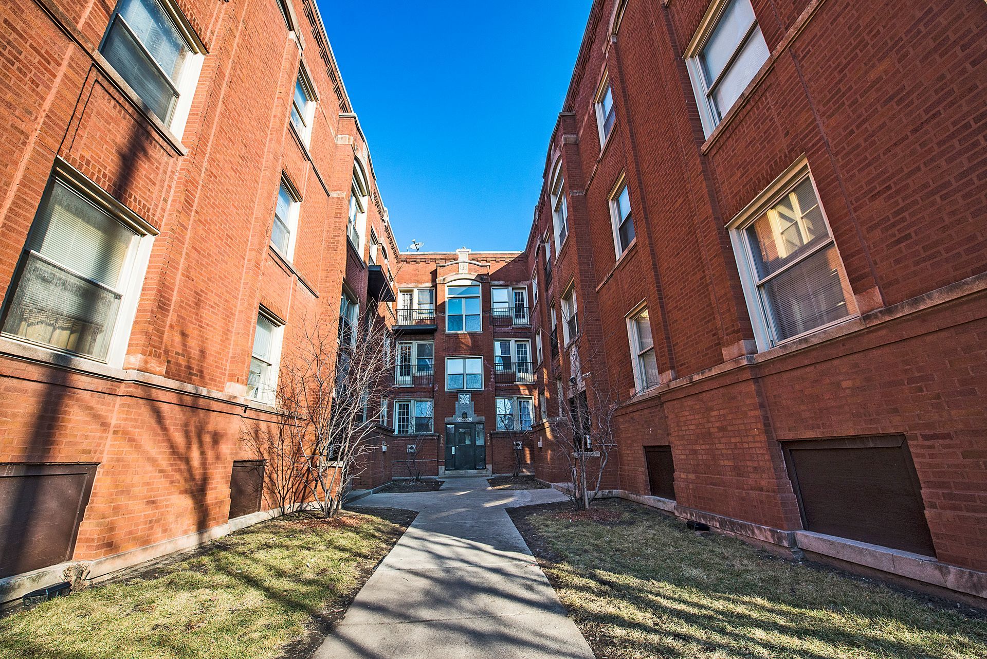 Red brick apartment buildings flank a walkway leading to a central entrance under a clear, blue sky.