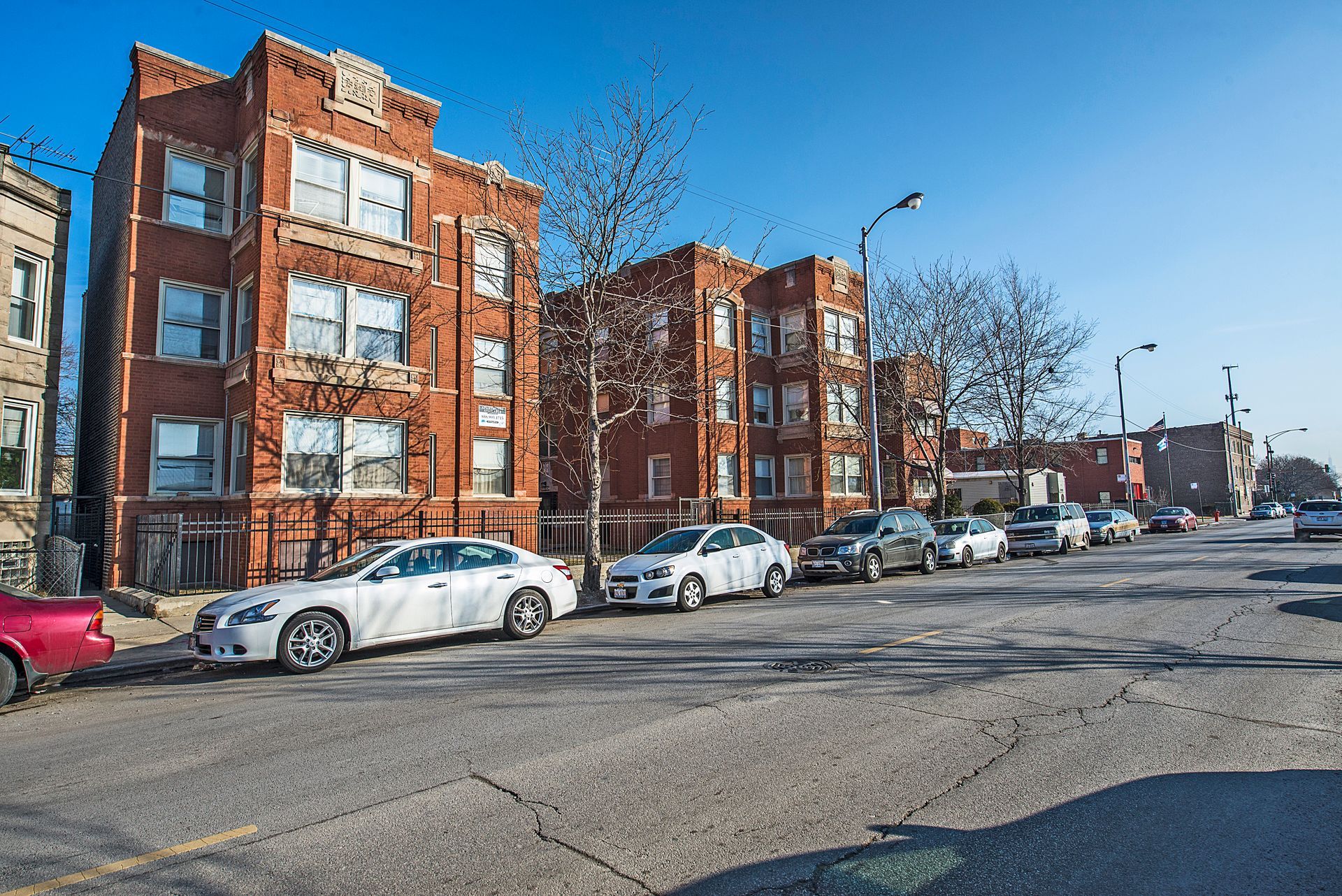 Row of red brick apartment buildings with cars parked on the street on a sunny day.
