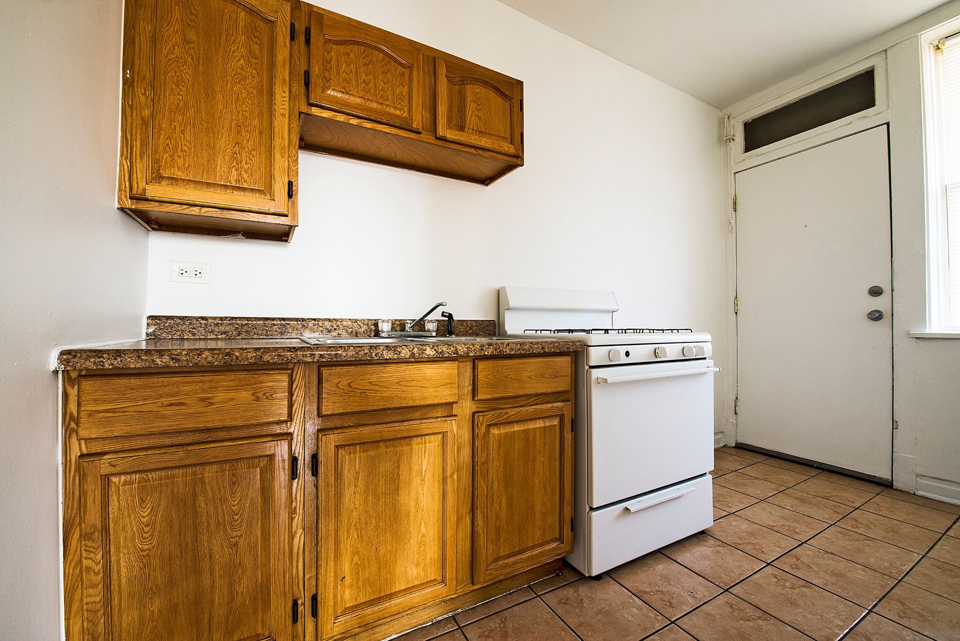 Small kitchen with wood cabinets, white stove, and tiled floor. A door and a window are present.