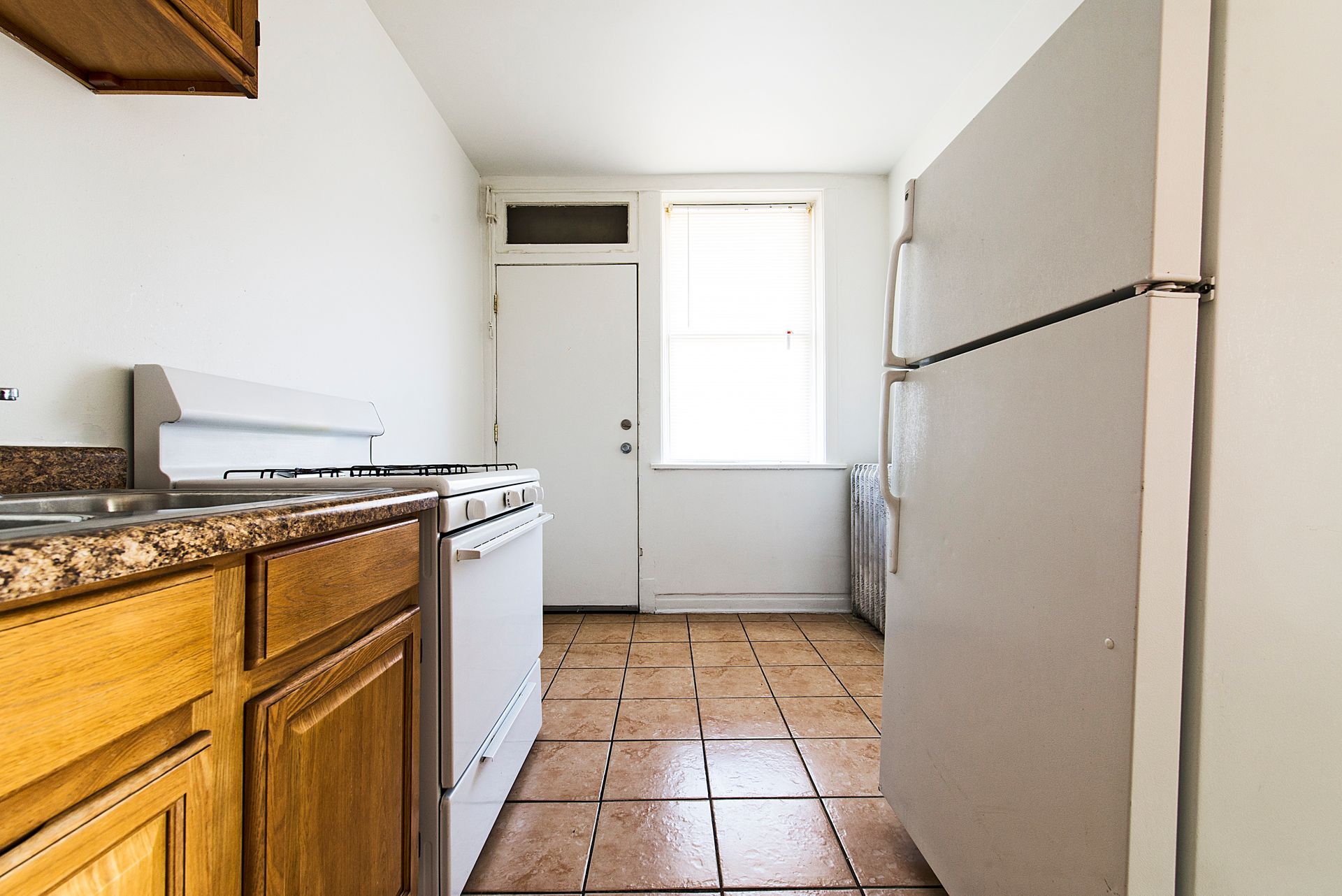 Kitchen with white appliances, wooden cabinets, and tile floor. Door and window at the end.