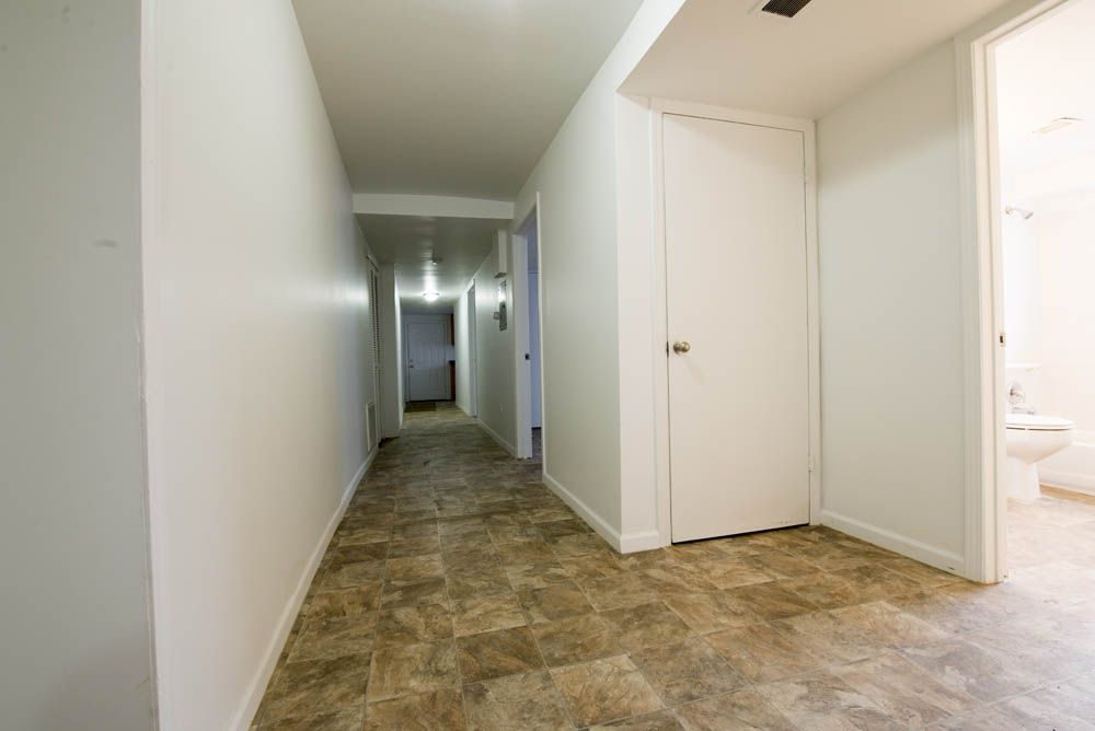 Narrow hallway with white walls and a brown tiled floor, leading to a doorway.