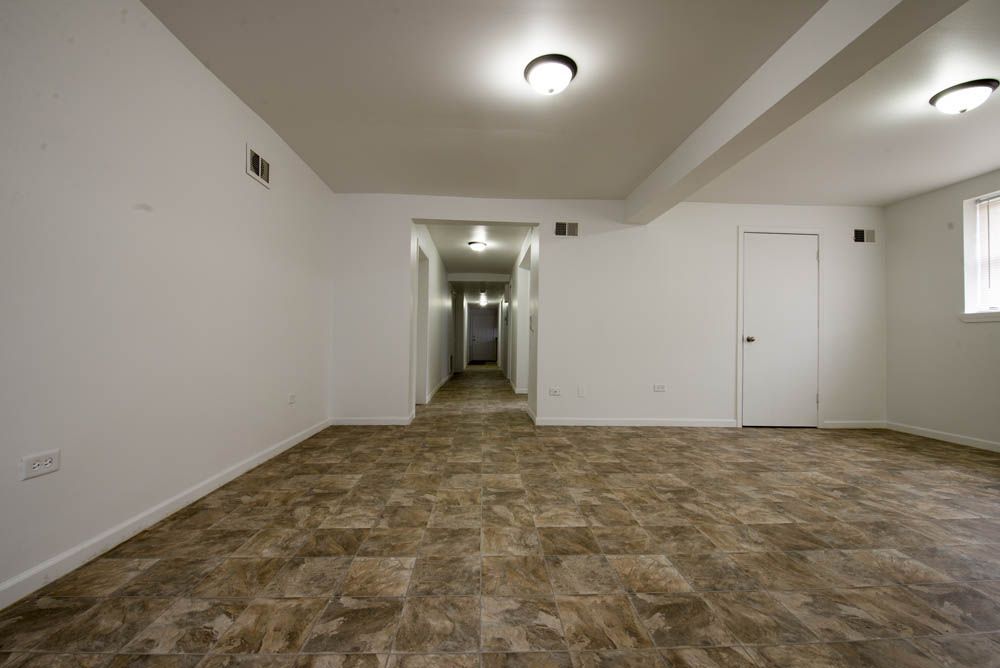 Empty interior room with beige flooring, white walls, and overhead lighting. A hallway leads to other rooms.