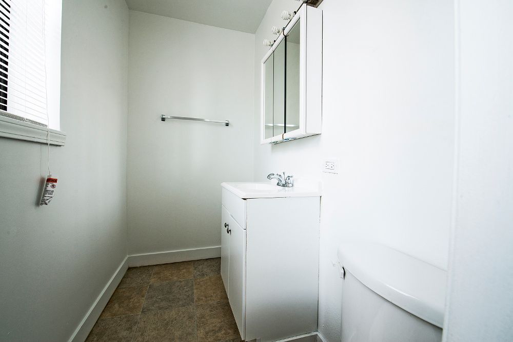 Small, white bathroom with vanity, toilet, and window with blinds.