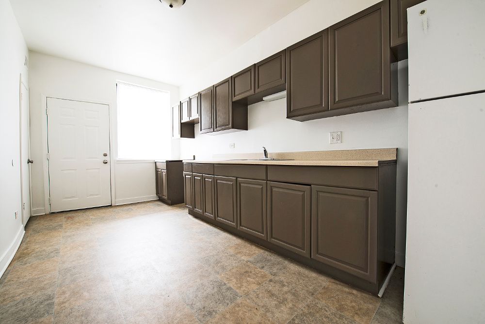 Empty kitchen with brown cabinets, a white refrigerator, and a door, with light coming in from a window.