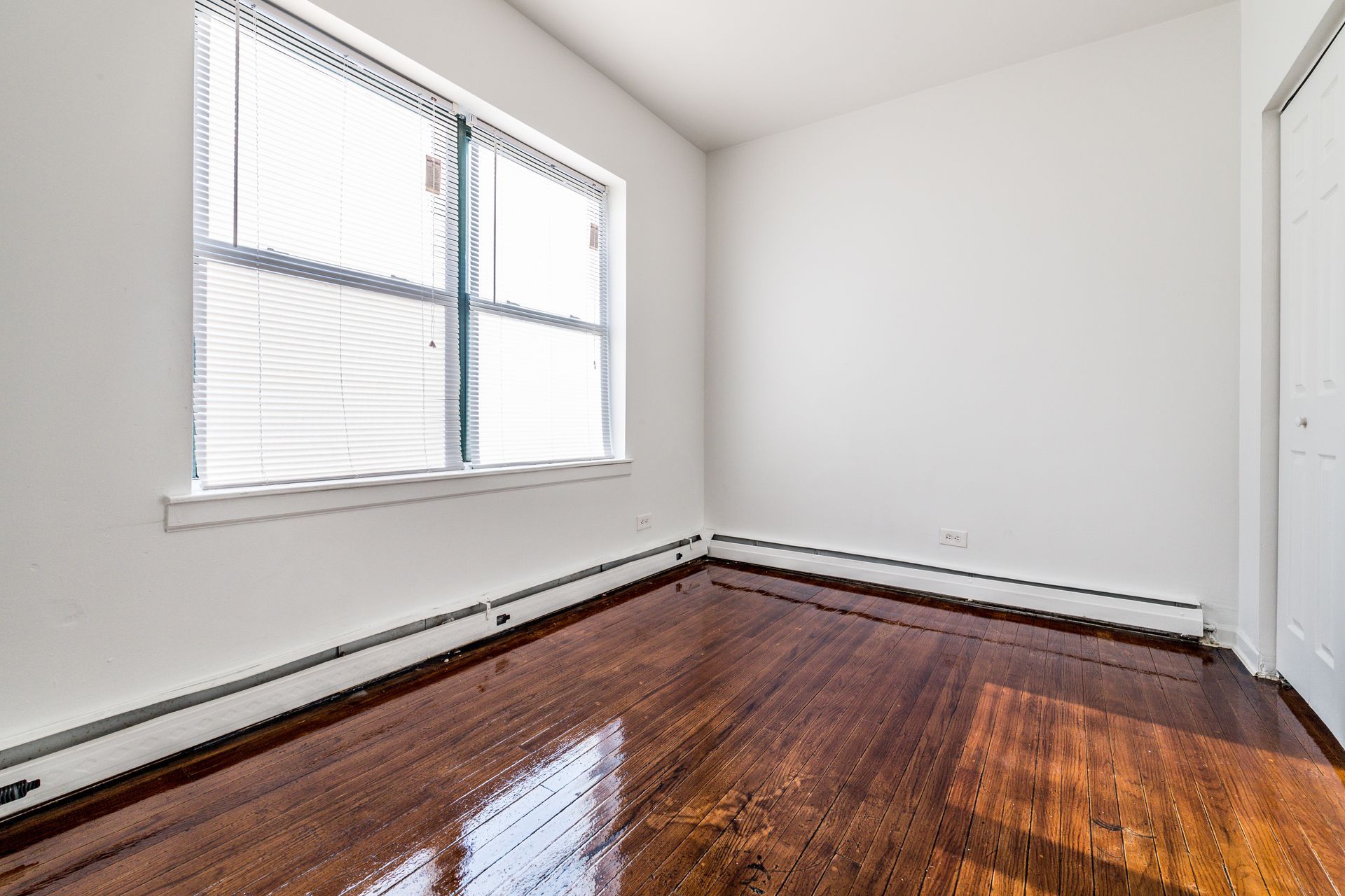 Empty room with a window, white walls, and shiny, dark wooden floors.