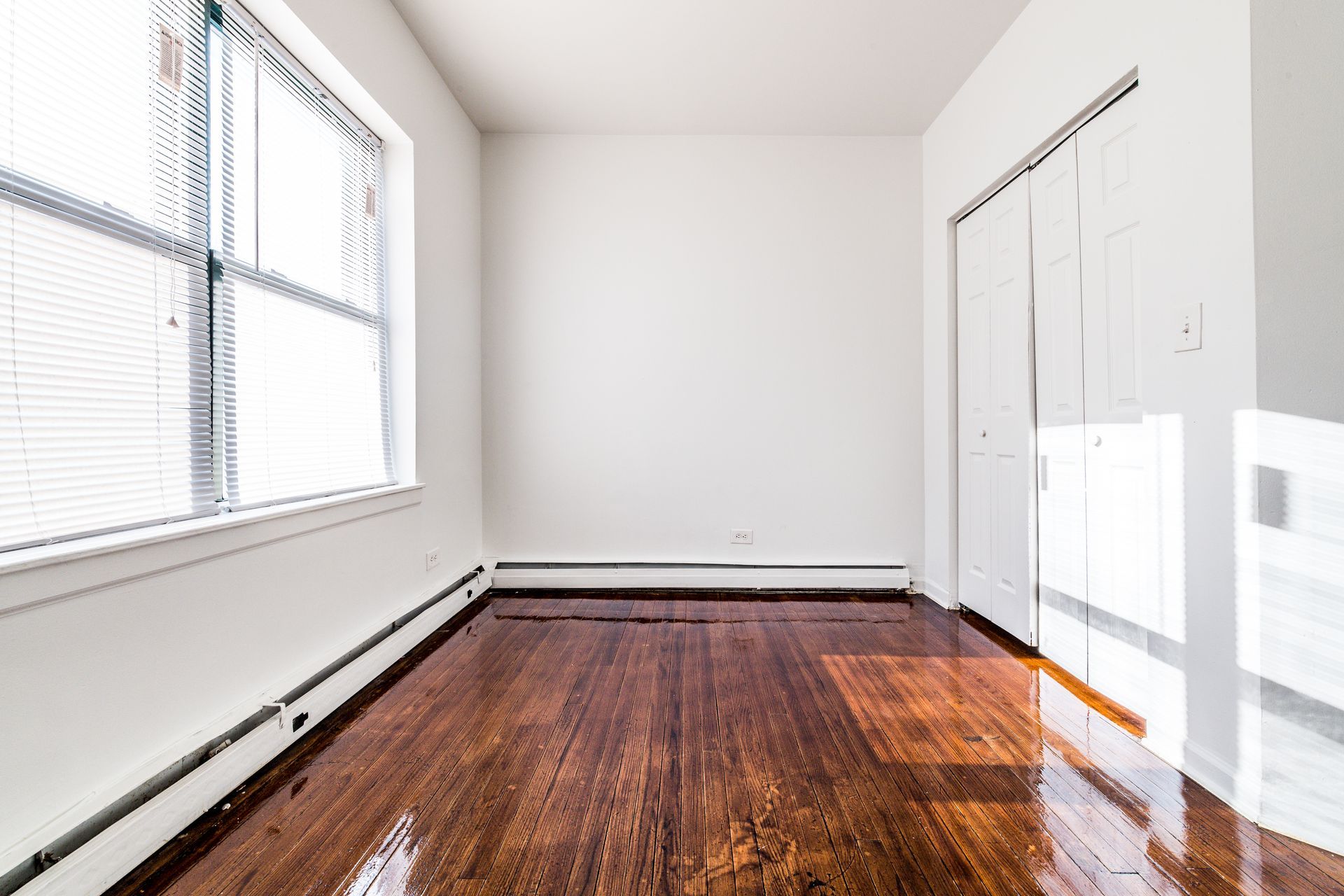 Empty room with wood floors, white walls, large window with blinds, and closet.