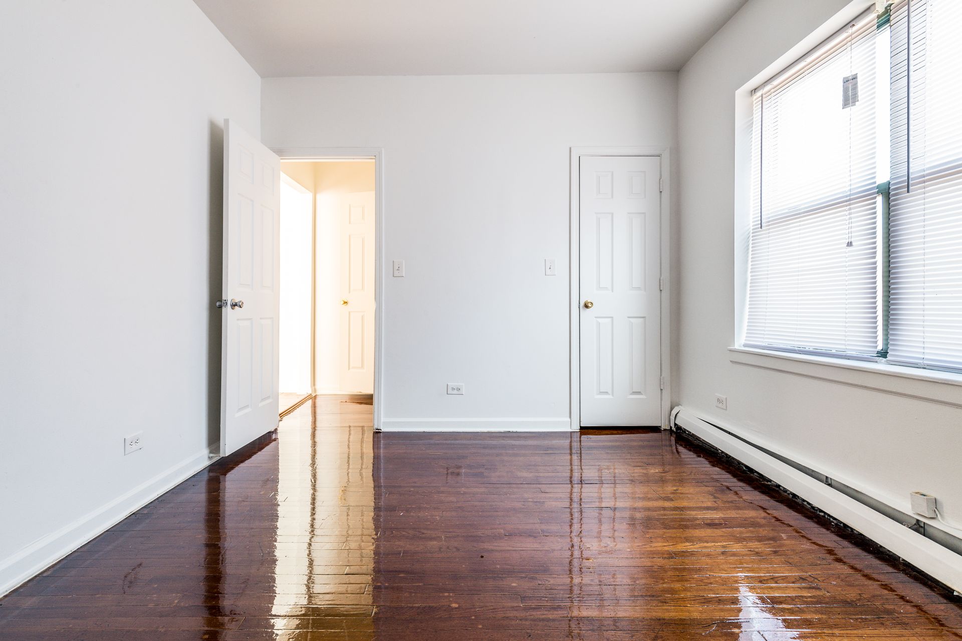 Empty room with hardwood floors, white walls, two closed doors, and a window.