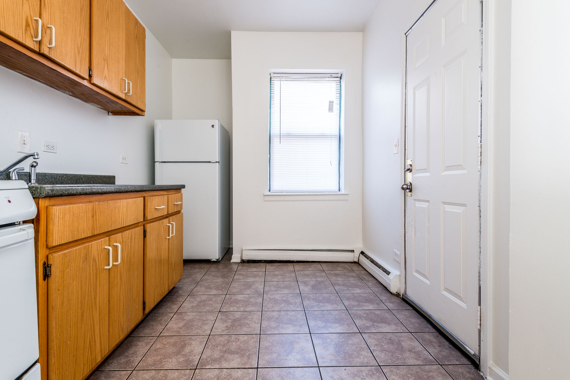 Small kitchen with wood cabinets, white appliances, window, and door. Tiled floor.