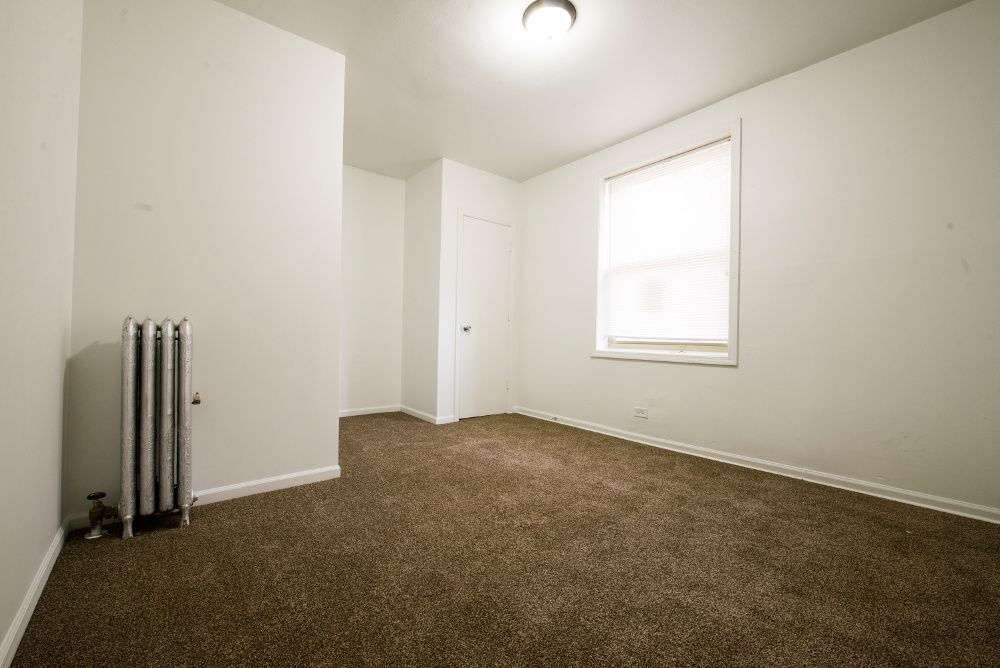 Empty room with brown carpet, white walls, radiator, window, and closed door.