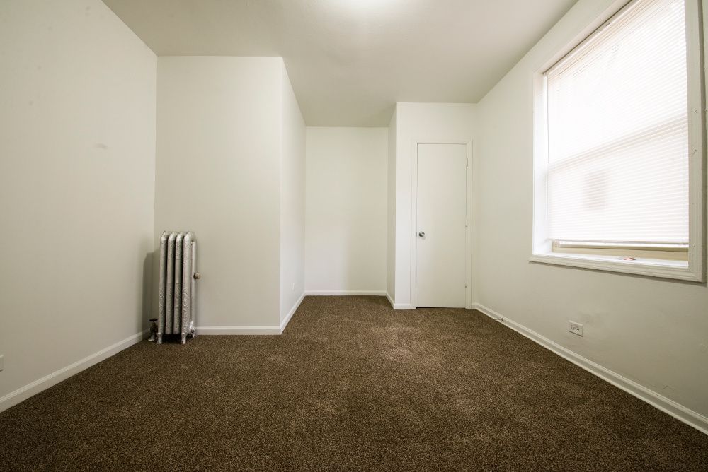 Empty room with brown carpet, white walls, radiator, window with blinds, and a closed door.