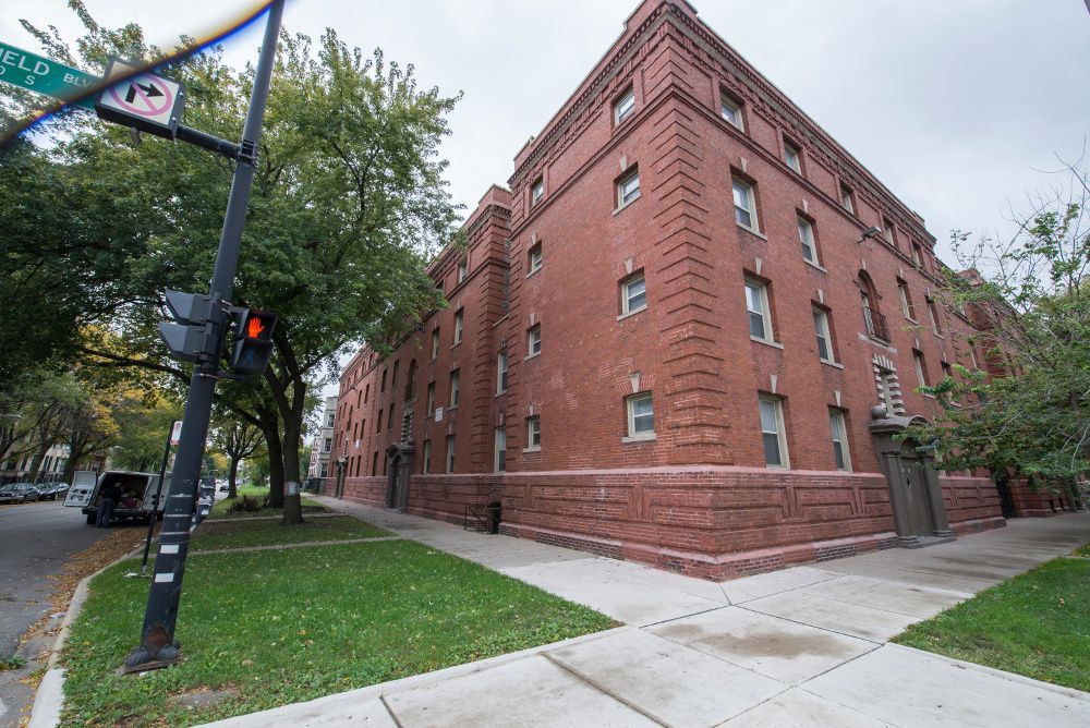 Brick apartment building on a street corner, with green lawn and trees.